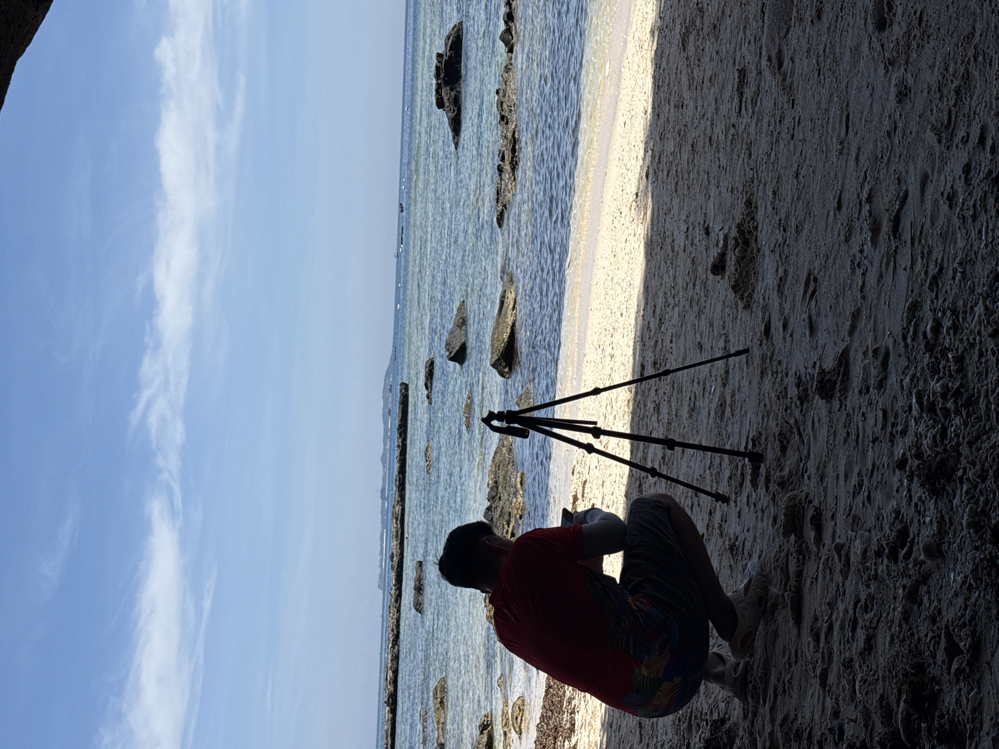 A person is crouching on a sandy beach, facing a tripod set up on the sand. The person is silhouetted against the bright water and sky, wearing a red shirt and patterned shorts. The sea is calm with scattered rocks, and the sky is blue with some wispy clouds. In the distance, there appears to be a shoreline with some structures or boats. The overall scene suggests a moment of photography or videography being prepared or in progress at a coastal location in the Lý Sơn Special Administrative Region of Vietnam.