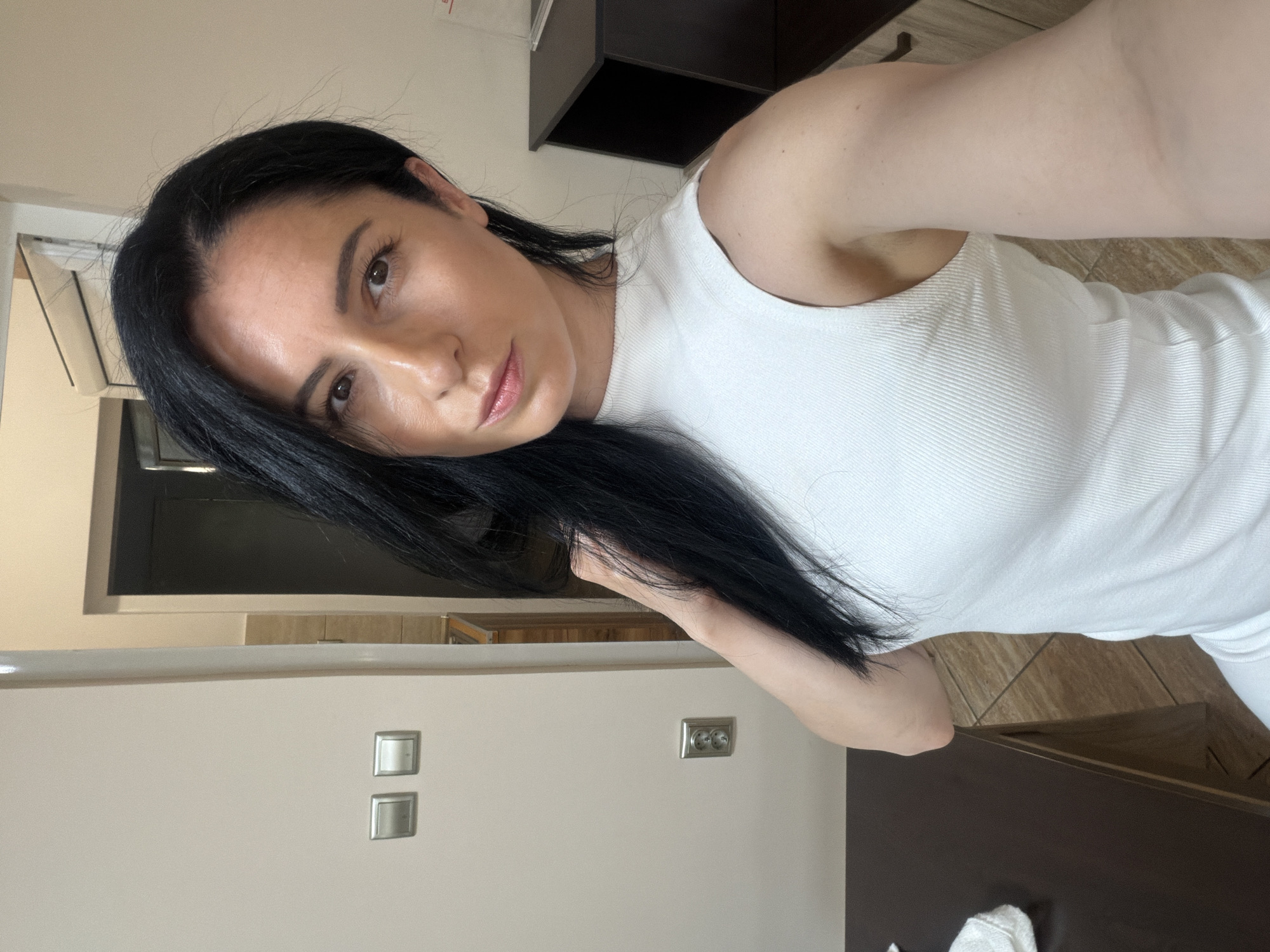 A woman with dark hair and fair skin is taking a selfie indoors. She is wearing a white tank top. In the background, there is a light beige wall with electrical outlets and light switches. A dark brown cabinet or shelving unit is also visible. The location cannot be determined as Pleven, Bulgaria.