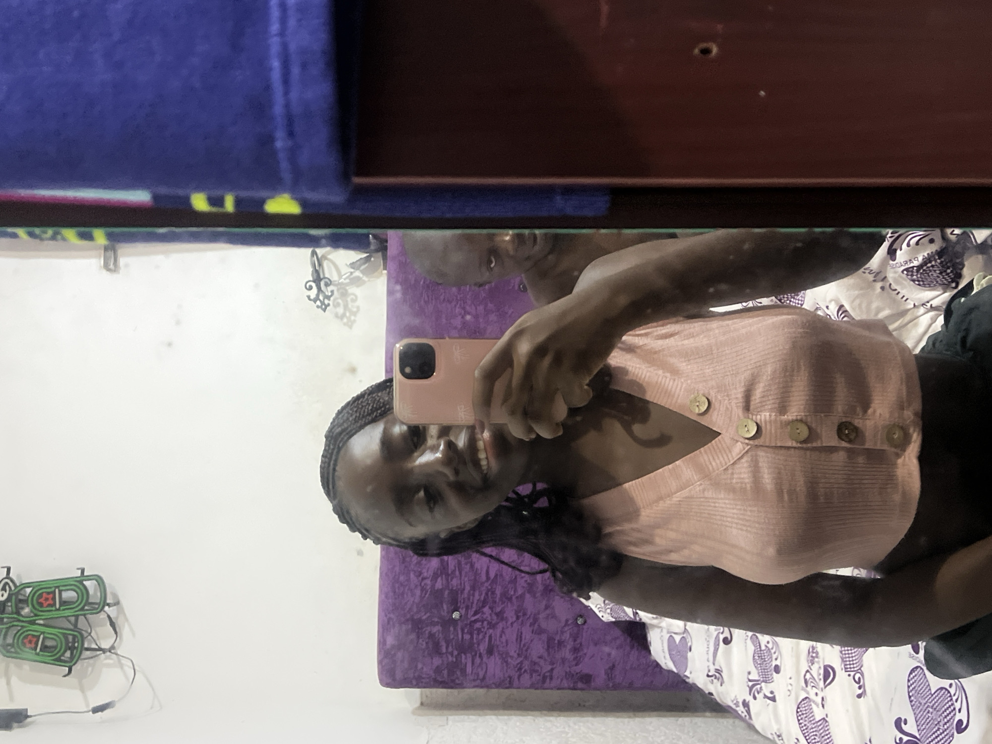 The image shows a young woman taking a selfie in a mirror. She is holding a rose-gold smartphone, and her reflection shows her wearing a light pink button-down top and braids. In the background, a man with a shaved head is visible in the mirror's reflection. A purple upholstered headboard and a patterned bedspread are also visible. On the wall to the left of the mirror, there is a neon green decorative light fixture. The location is likely an indoor space, potentially a bedroom, in an unknown city in Nigeria.