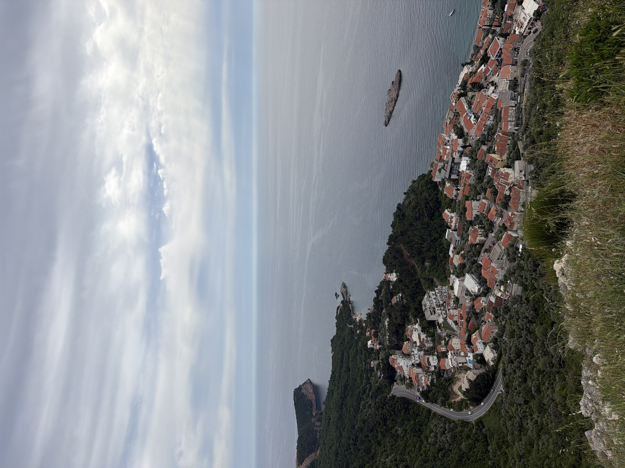 The image captures a view of the Montenegrin coast near Sveti Stefan. The scene encompasses a coastline dotted with buildings with red tile roofs, indicative of Mediterranean architecture, extending down to the shoreline where a winding road can be observed. The Adriatic sea stretches into the horizon under a sky with light cloud cover. A rock formation rises from the sea. Foreground elements include grass and rocky terrain.