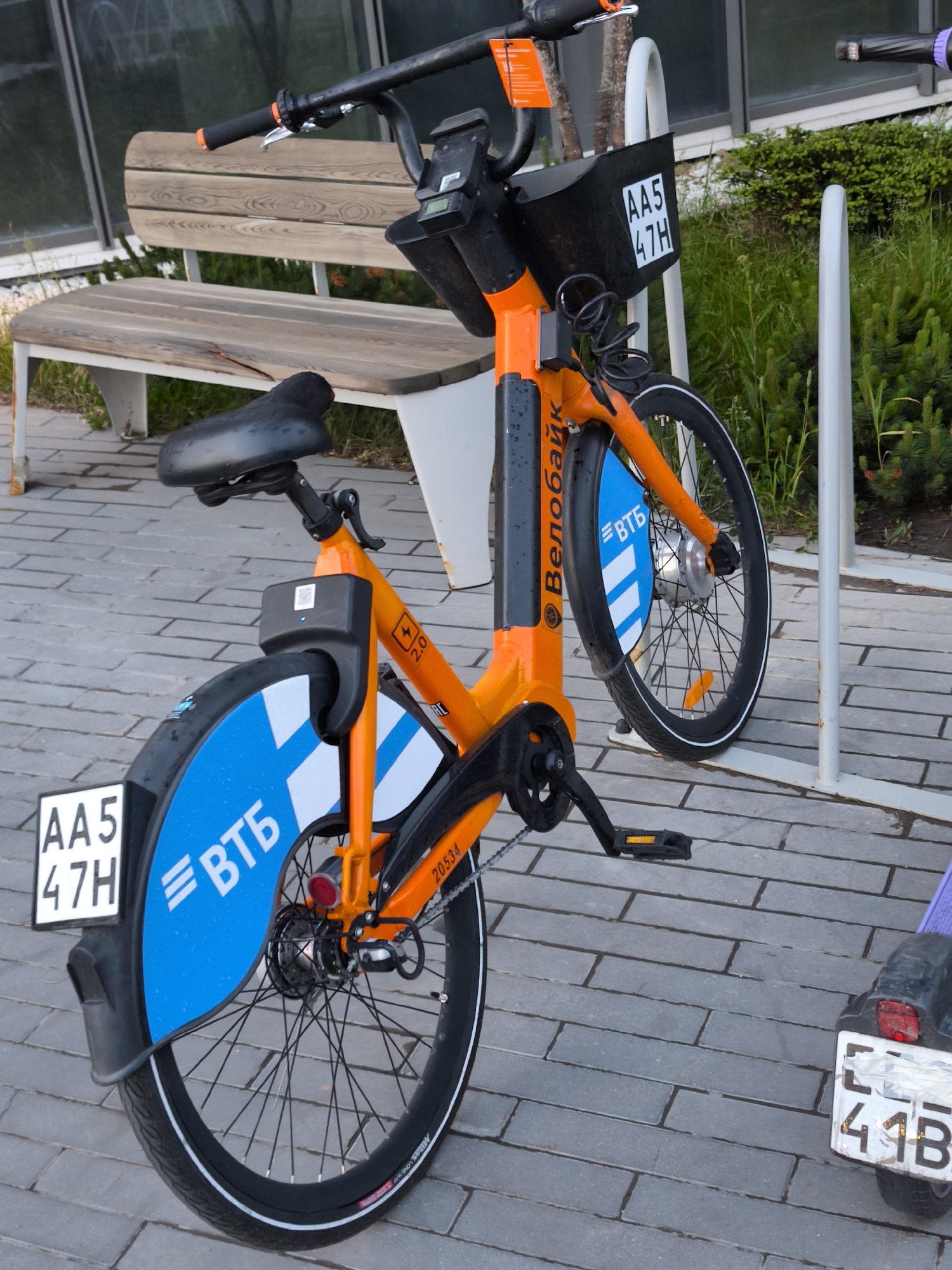 An orange Велобайк brand rental bicycle is parked on a paved brick surface in Moscow, Russia. The bike has a black seat and handlebars. It has a front basket and blue fender plates with the VTB logo. A license plate with the number "AA5 47H" is attached to both the front and rear fenders. A wooden bench with a white frame is visible in the background along with some greenery. The scooter parked on the right side of the bike has a partially-covered license plate showing "41B."