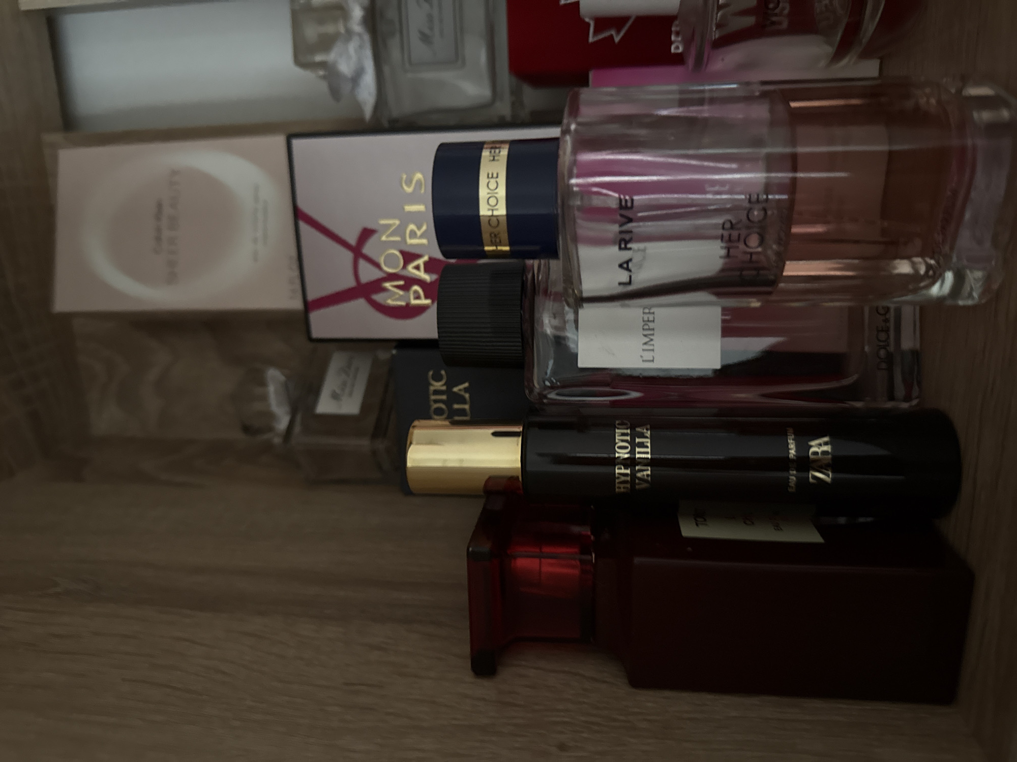 The image shows a shelf with several perfume bottles. The perfumes are arranged in a somewhat cluttered manner. In the foreground, there is a dark red bottle with a red cap on the left. To its right is a tall, dark bottle with a gold cap labeled "HYPNOTIC VANILLA" and "ZARA". Next to that is a clear bottle with a dark cap, and in front of it is a clear bottle with a blue and gold cap labeled "LA RIVE HER CHOICE". In the background, there are other perfume boxes and bottles. The shelf appears to be made of wood. The lighting suggests an indoor environment. There are no people visible in the image. The location is a shelf in a private residence in Chișinău, Moldova.