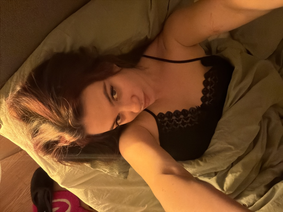 A woman lies on her left side in bed, partially covered by a light green comforter. She wears a black camisole with lace trim and gazes directly at the camera. Her hair is brown with reddish highlights. To the left, a pair of black slippers rest on a pink and white rug. The lighting is warm and muted, casting a soft glow across the scene. The bed appears to be against a gray headboard.