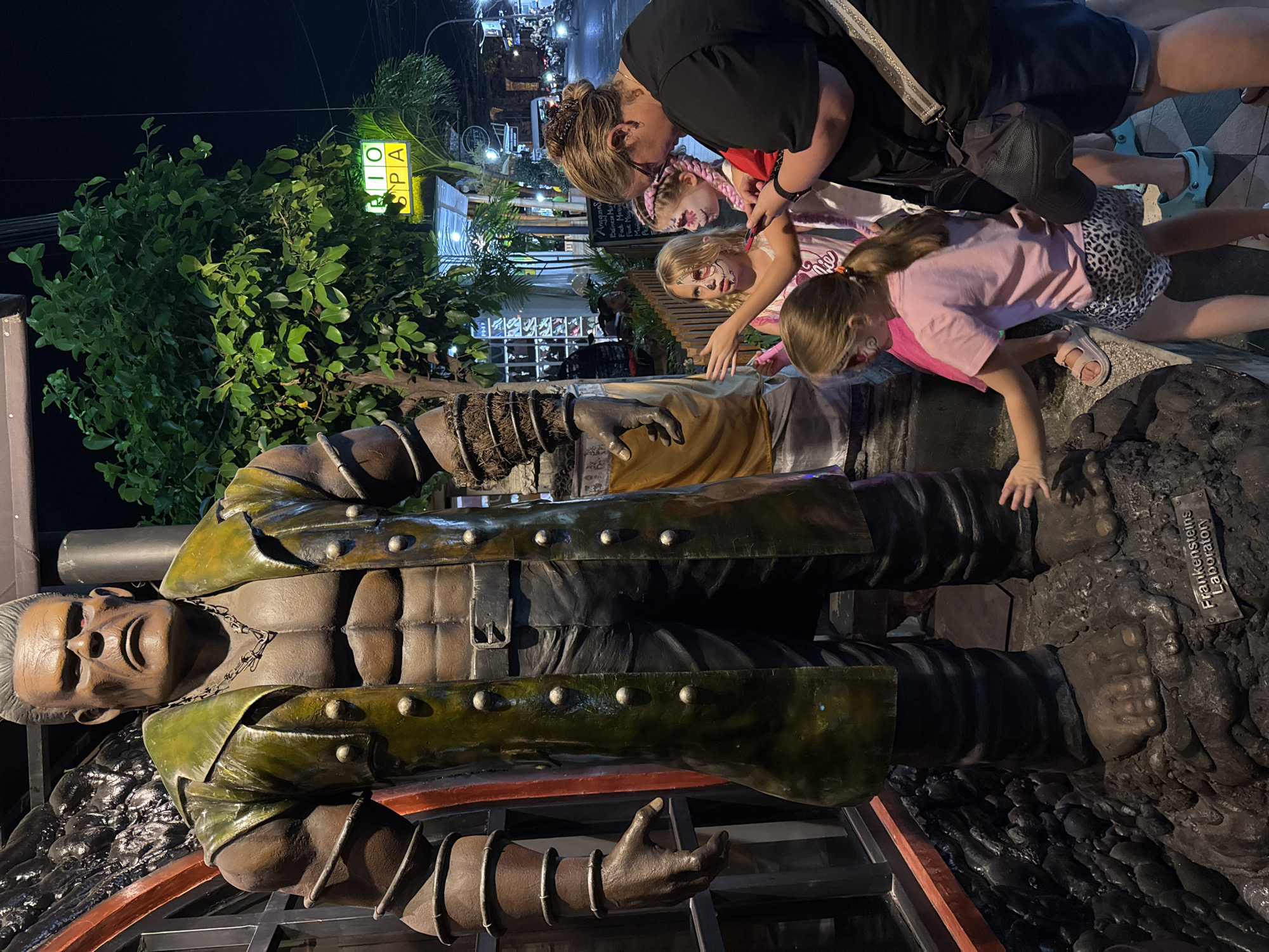 In Kuta, Indonesia, at night, a large, imposing statue of Frankenstein's monster stands on a rocky base, outside what appears to be a building labeled "Frankensteins Laboratory." The statue is dressed in a green trench coat and black pants. In front of the statue, a woman with blonde hair and glasses, wearing a black shirt and denim shorts, is with three young girls. Two of the girls have elaborate face paint resembling skulls or monsters. The girl closest to the statue, wearing a pink t-shirt and leopard print shorts, is reaching out to touch the base of the statue. The other two girls are looking at the statue, with one in a pink top and the other in a white top. Behind them, a street scene is visible with shops, palm trees, and motorcycles in the background. A neon green sign with "RIO SPA" is illuminated. The lighting suggests it is nighttime.