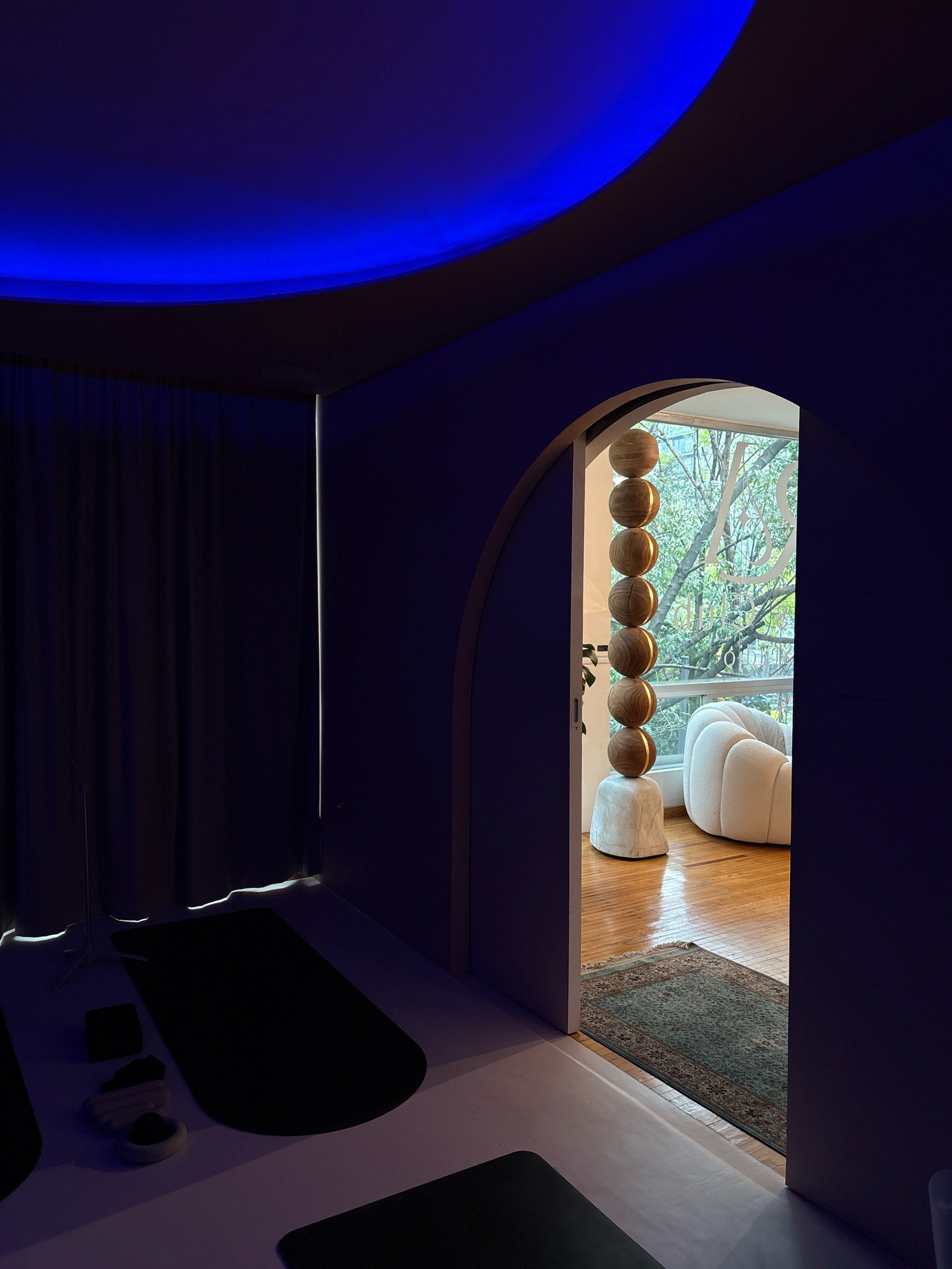 A dimly lit, modern room with a curved ceiling illuminated by vibrant blue LED lights. The room features dark purple walls and floor mats suggesting a yoga or meditation space. Through an arched doorway, a bright room is visible with polished wooden floors, a large window, and a white, puffy armchair. A decorative wooden sphere sculpture on a stone base stands near the doorway, and a patterned rug lies on the floor in the adjacent room. The overall ambiance is serene and contemporary. The location is a contemporary yoga studio in Roma Norte, Mexico City, Mexico.