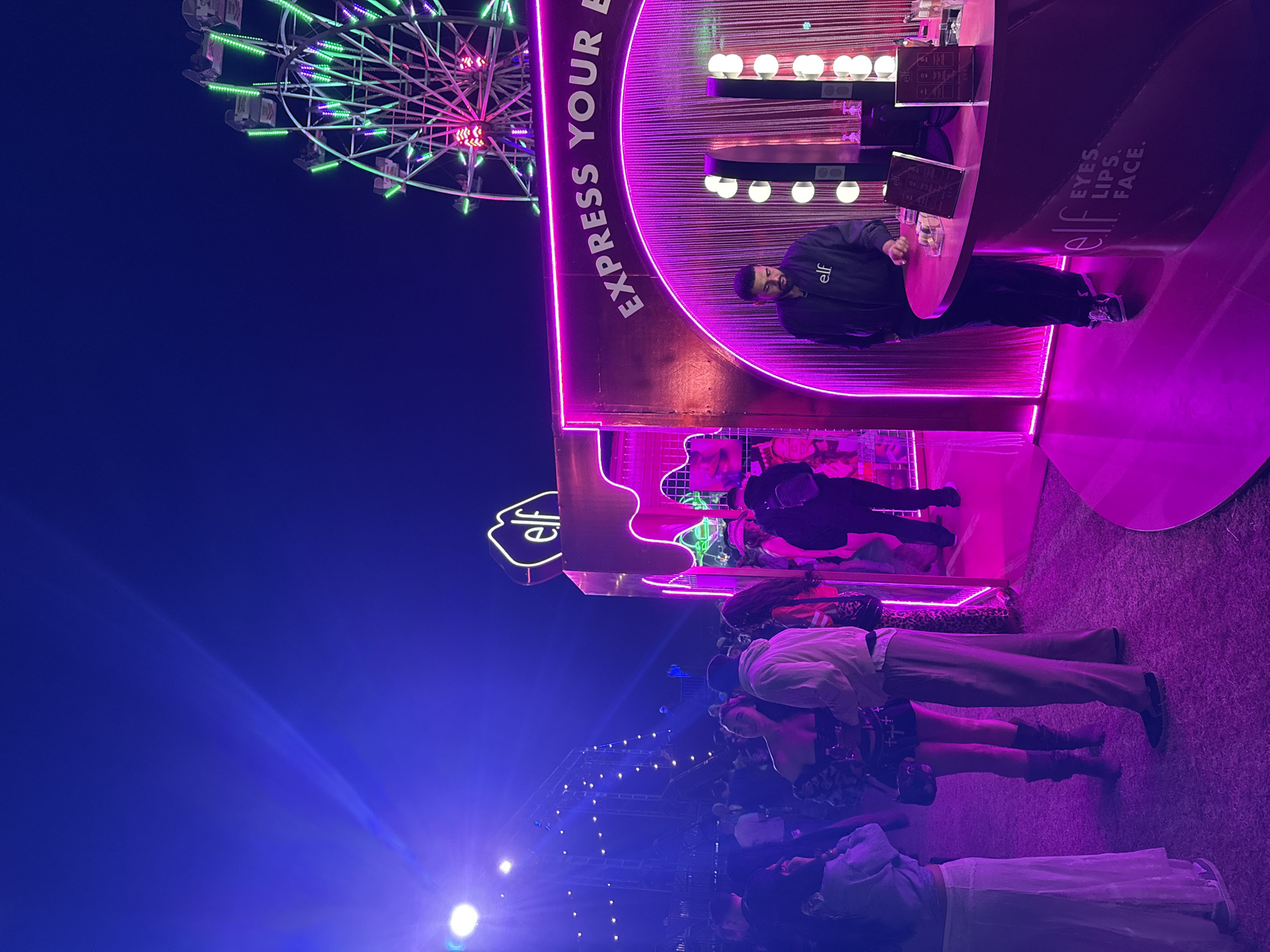 At night in Thermal, USA, an illuminated Ferris wheel looms near an E.L.F. booth. The booth, featuring a rounded design with the words "EXPRESS YOUR E" and "eyes. lips. face.", is lit with neon pink and purple lights. A man in a black jacket stands at a round counter inside the booth. People are gathered in front of the booth, partially obscured by a strong blue light. String lights are visible in the background.