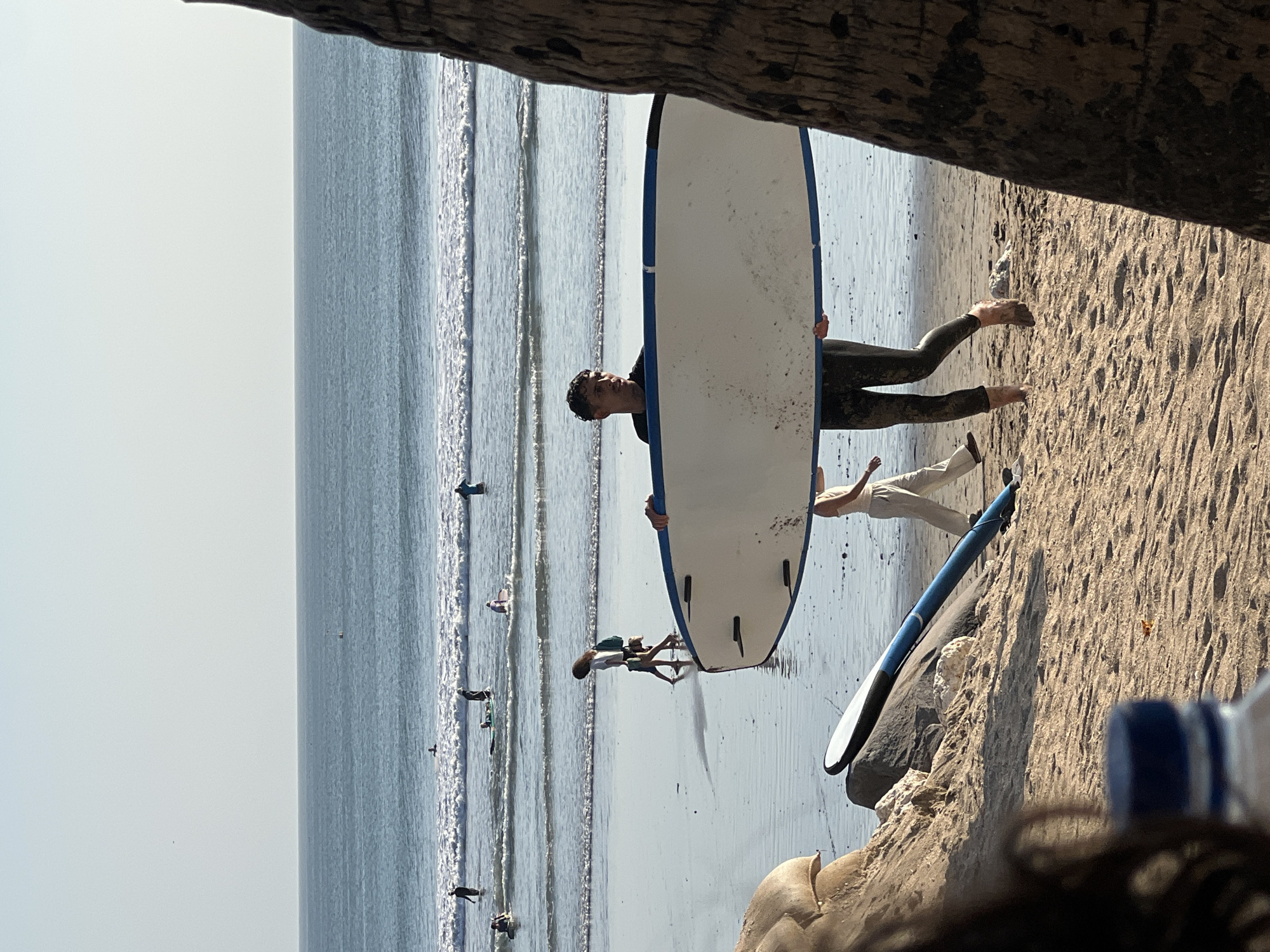 The image depicts a sunny day at Kuta Beach, Indonesia, a popular surfing destination. In the foreground, a person in a dark wetsuit stands holding a white surfboard with blue trim. They are walking on the wet sand near the water's edge. To the left of this individual, a blue surfboard leans against rocks. Further back, several other people are visible in the ocean, some surfing and others in the water. The waves are small to medium, indicating conditions suitable for beginners. The beach itself is sandy, with some rocks and vegetation visible in the background. The sky is clear and bright, typical of a tropical climate. The overall atmosphere is one of leisure and outdoor activity.