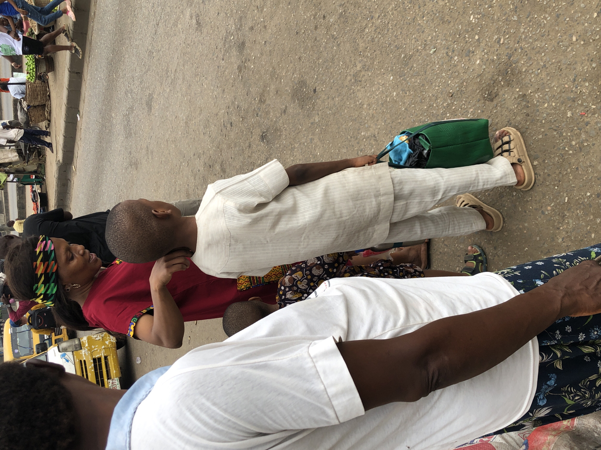 In Shangisha, Nigeria, a street scene unfolds with several individuals and a yellow vehicle. In the foreground, a person wearing a white shirt and blue floral skirt is partially visible from the side. To their right, a young boy in a cream-colored traditional outfit stands with his back to the camera, carrying a green bag. Beside him, a woman with a vibrant, colorful headband and a red dress is interacting with someone behind her. In the background, a yellow bus is parked on the street, and other people are scattered around, suggesting a busy urban environment. The ground is paved, and market stalls or vendors are present on the side of the road.