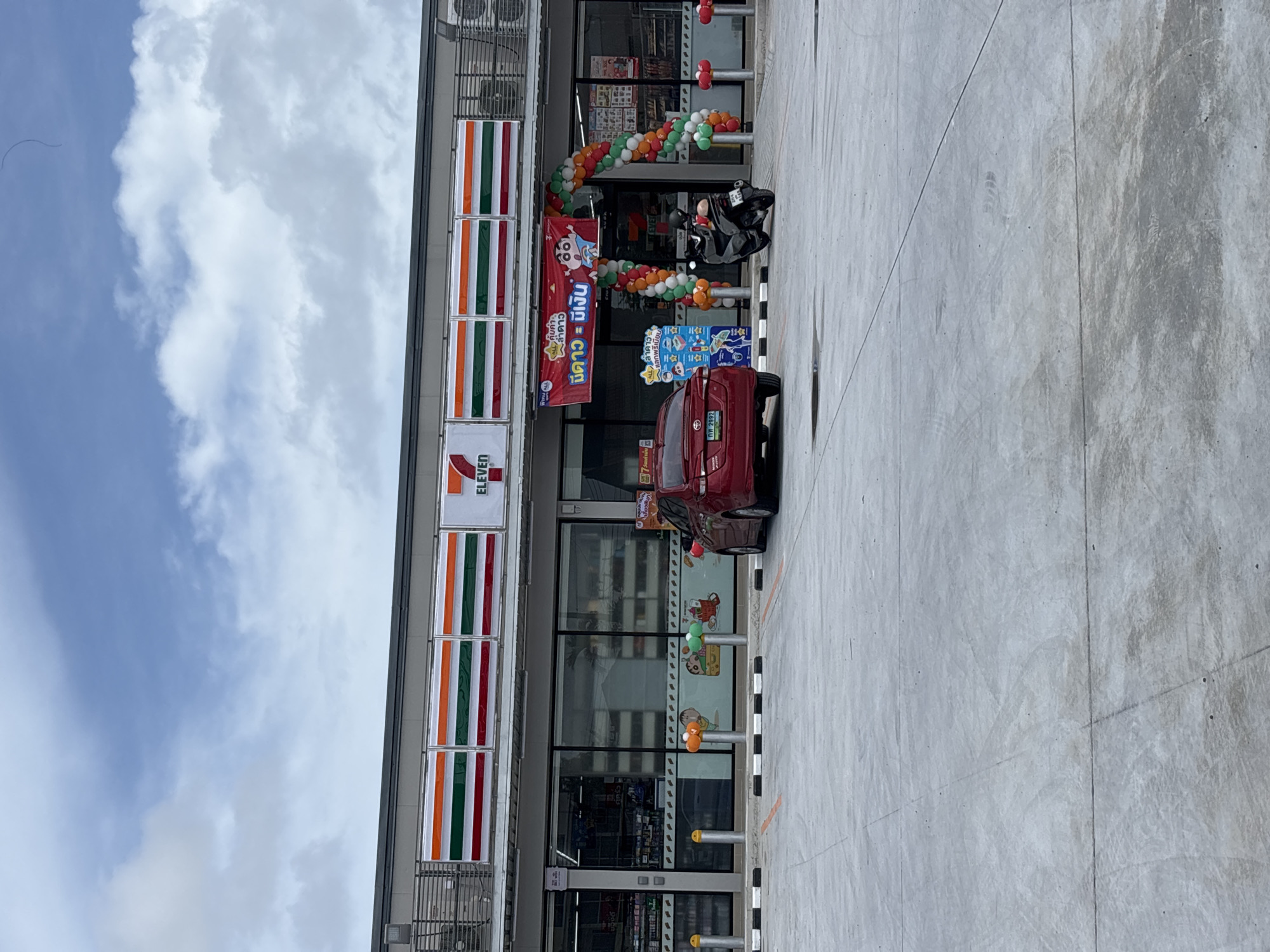 A 7-Eleven convenience store is depicted in Rawai, Thailand. The store has a prominent facade with the 7-Eleven logo and horizontal stripes in orange, white, and green. A large archway made of colorful balloons is positioned at the entrance, with a banner above it displaying Thai text and a cartoon character. Two vehicles, a red car and a black motorcycle, are parked in the foreground on a concrete lot. The lot is marked with parking lines and bollards. The sky above is partly cloudy.