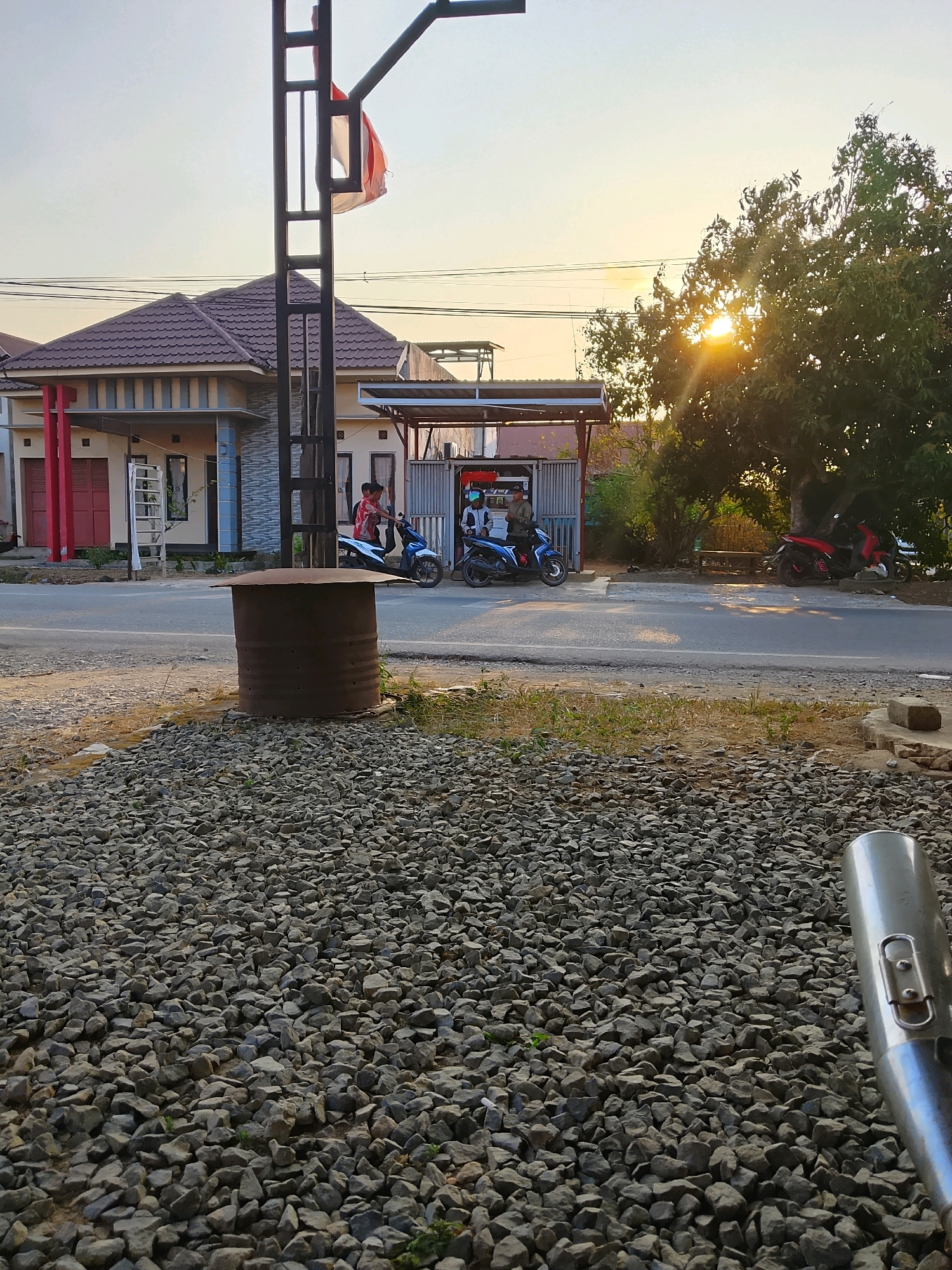 The image depicts a street scene in Pelaihari, Indonesia, during what appears to be late afternoon or early evening, given the warm, low angle of the sun. The foreground is dominated by a densely packed area of grey gravel. To the left, a large, rusty metal barrel with a lid sits on the gravel.
In the middle ground, a paved road runs horizontally across the frame. On the left side of the road, a modern-looking house with a tiled, brown roof and light-colored walls is visible, featuring red doors and white window frames. A ladder leans against the side of the house. Further down the road, across from the house, is a small roadside structure with a light-colored roof, possibly a stall or shop. Two young men are present at this structure. One is on a blue motorcycle, and another is standing nearby, appearing to interact with someone inside the structure.
To the right of the middle ground, several trees are present. The largest is a mature tree with dense green foliage, through which the setting sun casts rays of light, creating a lens flare effect. Parked near this tree are two motorcycles, one red and one dark blue, with a person standing near the red one.
The sky is a pale, warm hue, indicative of the time of day. Overhead, power lines are strung across the scene. A tall, black metal pole with a red and white flag attached to it stands prominently in the left-center of the image, interrupting the view of the house and the sky. The overall atmosphere is tranquil and rural, with elements of modern housing and everyday street life.