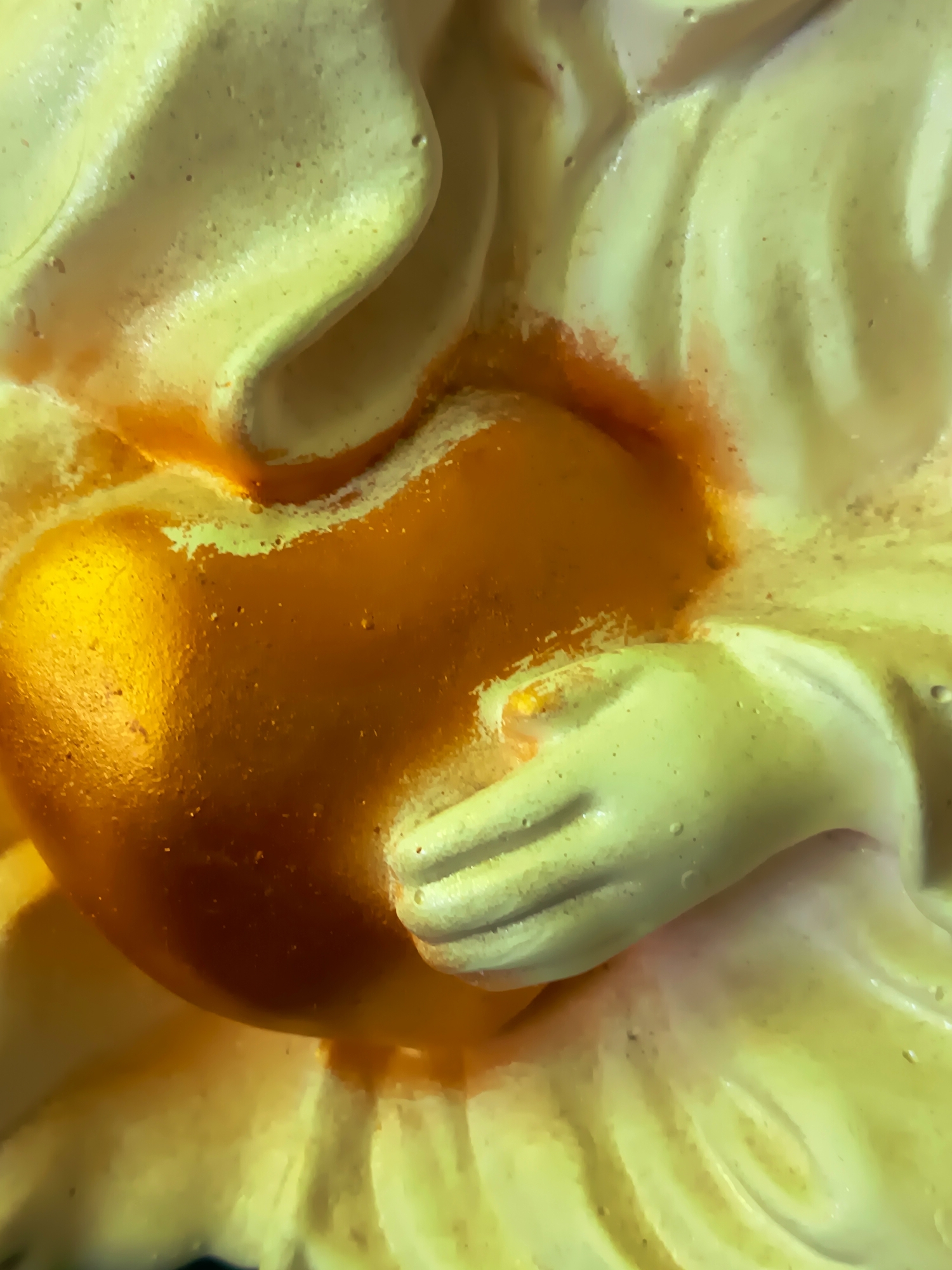 This is a macro photograph of a religious statue, possibly depicting a depiction of Christ or a saint, holding a golden heart. The statue is rendered in a pale, creamy material with accents of orange and gold. The texture of the material appears slightly porous. The lighting is warm and highlights the reflective quality of the golden heart, creating a luminous effect. The background is out of focus and appears to be a continuation of the same material or a similar textured surface. The image is very close-up, focusing on the hands and the heart, with minimal environmental context visible. The specific location in Izmail, Ukraine, is not discernible from the image itself.