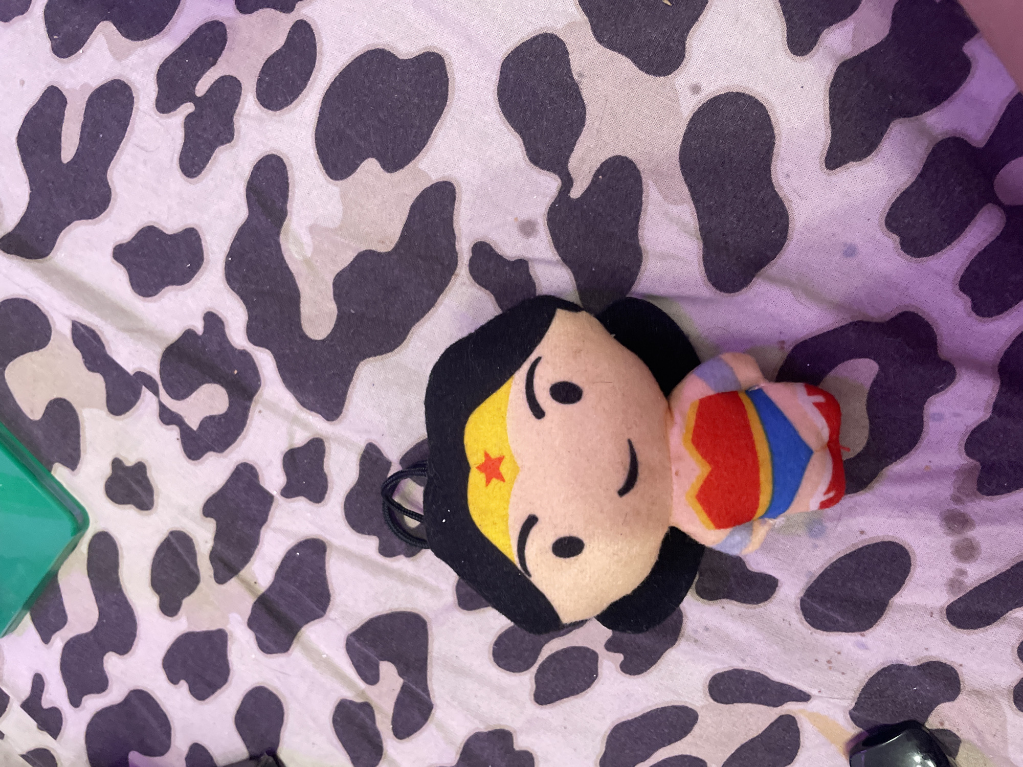 A plush toy of Wonder Woman is lying on a white and black spotted fabric. The toy is positioned in the center of the frame, facing forward. It has black hair, a yellow tiara with a red star, and a smiling face. Its outfit consists of a red top with a gold trim, a blue bottom, and red and white striped boots. The fabric underneath appears to be a blanket or sheet with a cow-print pattern. In the upper left corner of the image, a portion of a green object is visible. The lighting suggests indoor ambient light with some purple hue. The image is a close-up shot, with the toy being the primary subject. The location in the city of Kokkorevo, Russia is not evident from the image.