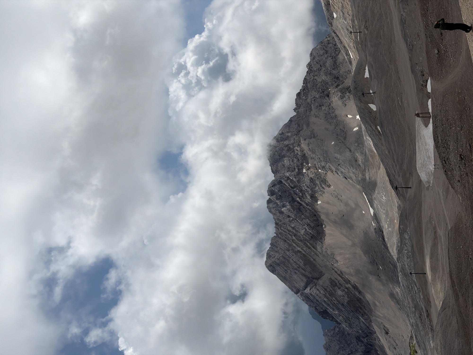 The image depicts a vast, desolate mountain landscape under a dramatic sky filled with white and grey clouds. The foreground is a barren, rocky terrain with several poles, remnants of what appear to be ski lift infrastructure, scattered across the scene. Patches of snow are visible on the slopes. In the mid-ground, a rugged, imposing mountain range dominates, its rocky slopes showing signs of erosion and some lingering snow. The sky above is a mixture of bright blue patches and dense cloud cover, creating a sense of dynamic weather. In the lower right corner of the image, a lone figure wearing a hooded jacket is seen from behind, looking towards the mountains, their posture suggesting contemplation or observation. This scene is likely the Zugspitze, Germany's highest peak, located near Garmisch-Partenkirchen.