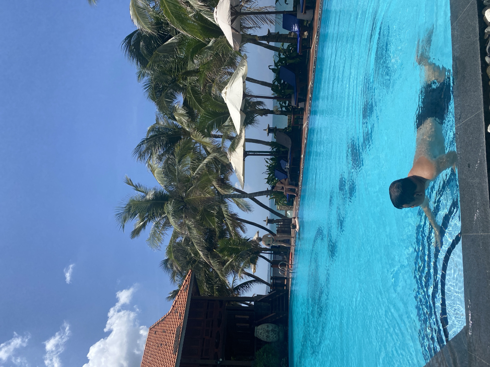 This image captures an outdoor scene at a resort or hotel with a swimming pool, ocean view, and tropical vegetation in Phu Thuy Ward, Vietnam, under a bright blue sky with sparse white cumulus clouds.
In the foreground, a man with short, dark hair is partially submerged in the vibrant turquoise-blue swimming pool, facing away from the viewer and slightly to the right. He is wearing dark swim shorts, and his upper back and shoulders are visible above the water. His left arm is extended forward, appearing to propel him along the water's surface. The pool's edge, made of dark grey/black textured tiles, is prominent in the immediate foreground. The pool water is clear, revealing patterns on the bottom tiles and sunlight reflections.
In the midground, the swimming pool extends towards a resort area. To the left, a traditional multi-story wooden building with a reddish-brown tiled roof stands beside the pool. An ornate, large, light-colored ceramic urn is visible near its base. Adjacent to the building, a metal pool ladder extends into the water. A person with light-colored hair, wearing a light-colored two-piece swimsuit, stands at the pool's edge near the ladder, facing away from the viewer, looking towards the ocean. Another person is partially submerged in the water nearby.
Further to the right, along the pool's edge, several lounge chairs with blue cushions are arranged under three large, light-colored parasols, providing shade. Two individuals are seated on lounge chairs. Beyond the parasols and lounge chairs, numerous tall palm trees with green fronds line the area, creating a tropical backdrop. Between the trees, a hazy light blue expanse of the ocean is visible, marking the horizon. A small decorative stone lantern is placed among the foliage near the ocean view.