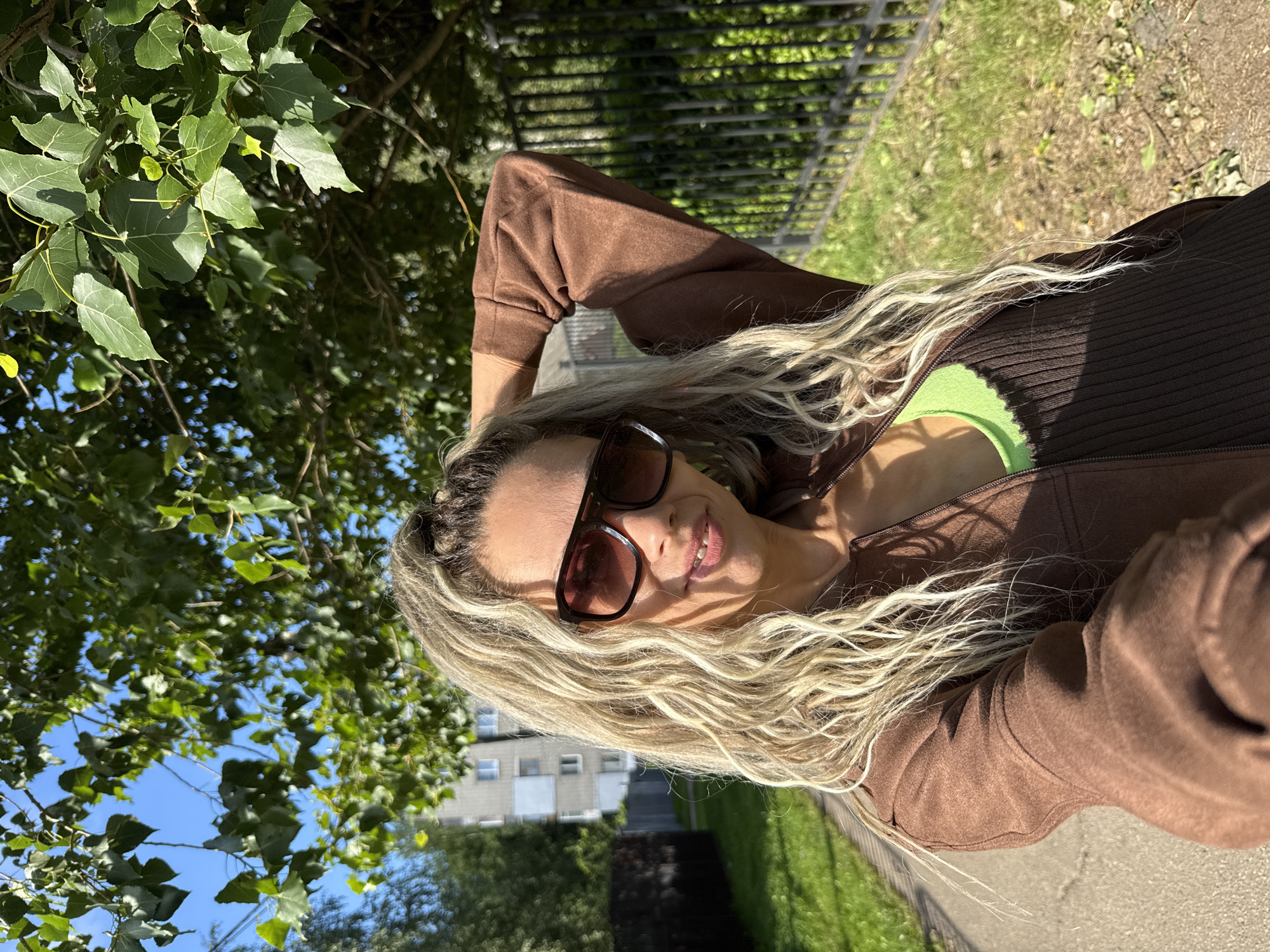 A woman with long, blonde, curly hair is taking a selfie outdoors. She is wearing large, dark-rimmed sunglasses, a brown zip-up jacket over a green ribbed tank top. Her left arm is raised and bent, with her hand behind her head. The background shows a paved walkway, a grassy area, a black metal fence, and a multi-story building. Trees with green leaves are visible overhead, with a clear blue sky. The setting appears to be a residential area in Rusanovo, Russia.