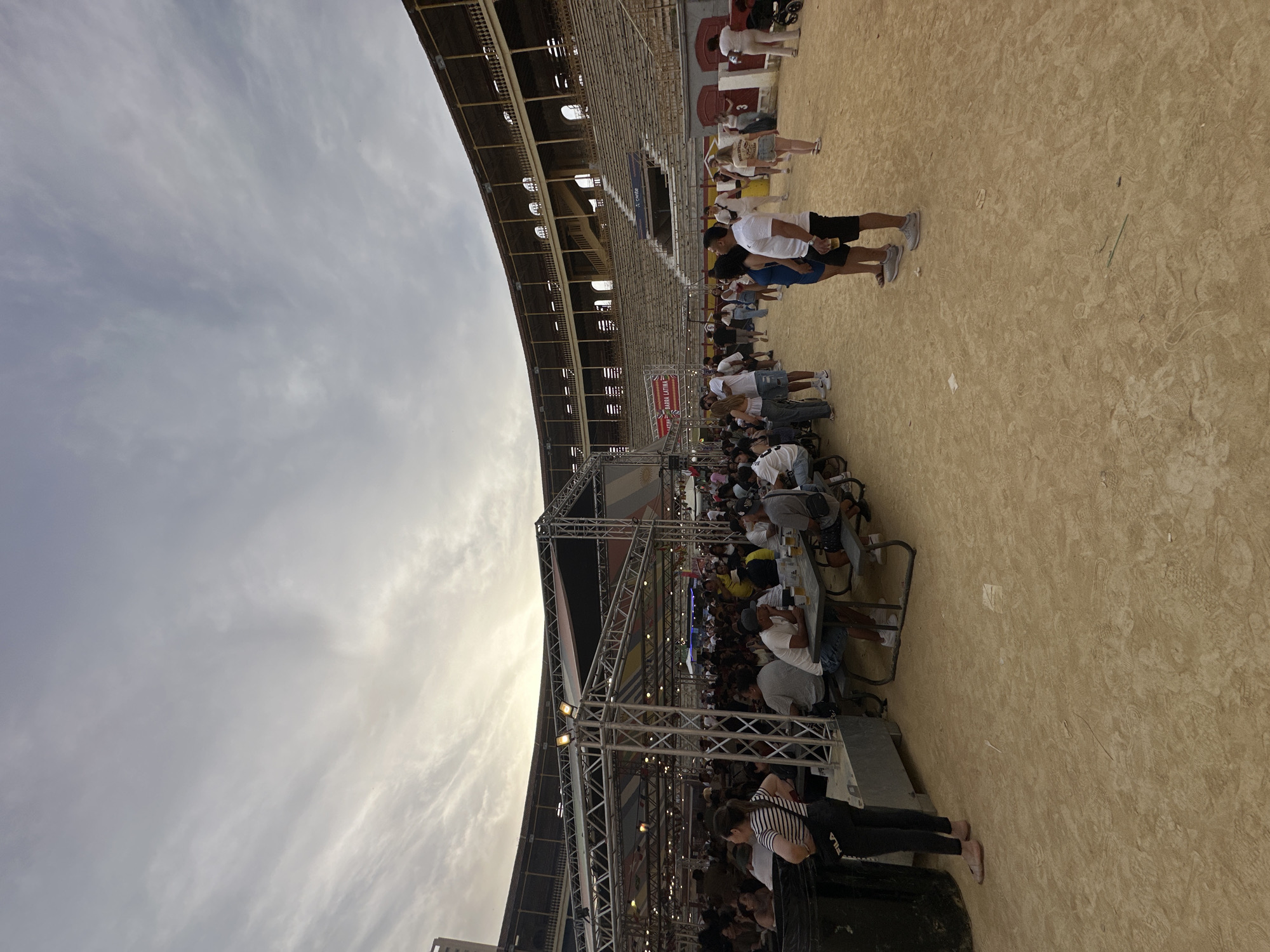 The image depicts an outdoor event taking place on the sandy arena floor of a bullring. The bullring's tiered, empty spectator stands, constructed with light-colored stone and featuring archways and balustrades, encircle the perimeter under an overcast sky.
In the foreground and midground, a large, temporary event structure made of silver metal trussing supports a dark canopy, likely providing shade or cover. Numerous string lights are suspended underneath this canopy. Various banners and flags with diverse patterns are draped from the trusswork, with one partially visible banner reading "BARRA LATINA."
A substantial crowd of individuals, primarily adults, occupies the arena floor. Many are gathered under and around the truss structure, where multiple picnic-style tables with attached benches are set up. People are seated at these tables, engaged in social interaction, with several visible holding or having glasses of beer. Other attendees stand and mingle both within the covered area and on the open sandy ground. Clothing is predominantly casual, suited for warm weather, including t-shirts, shorts, and dresses.
On the far left, a woman in a striped shirt and dark pants, bearing a "FILA" logo, stands next to a large, black industrial-style bin. Further right, on the open sand, a man and a woman stand together, the man holding a drink. In the background on the right, near the bullring wall, a woman in light attire is seen pushing a baby stroller. The sandy ground is visibly marked with numerous footprints and scattered small debris, including a green stick or straw. The sky is largely covered with thin, high clouds, indicating diffuse light, possibly late afternoon or early evening.
