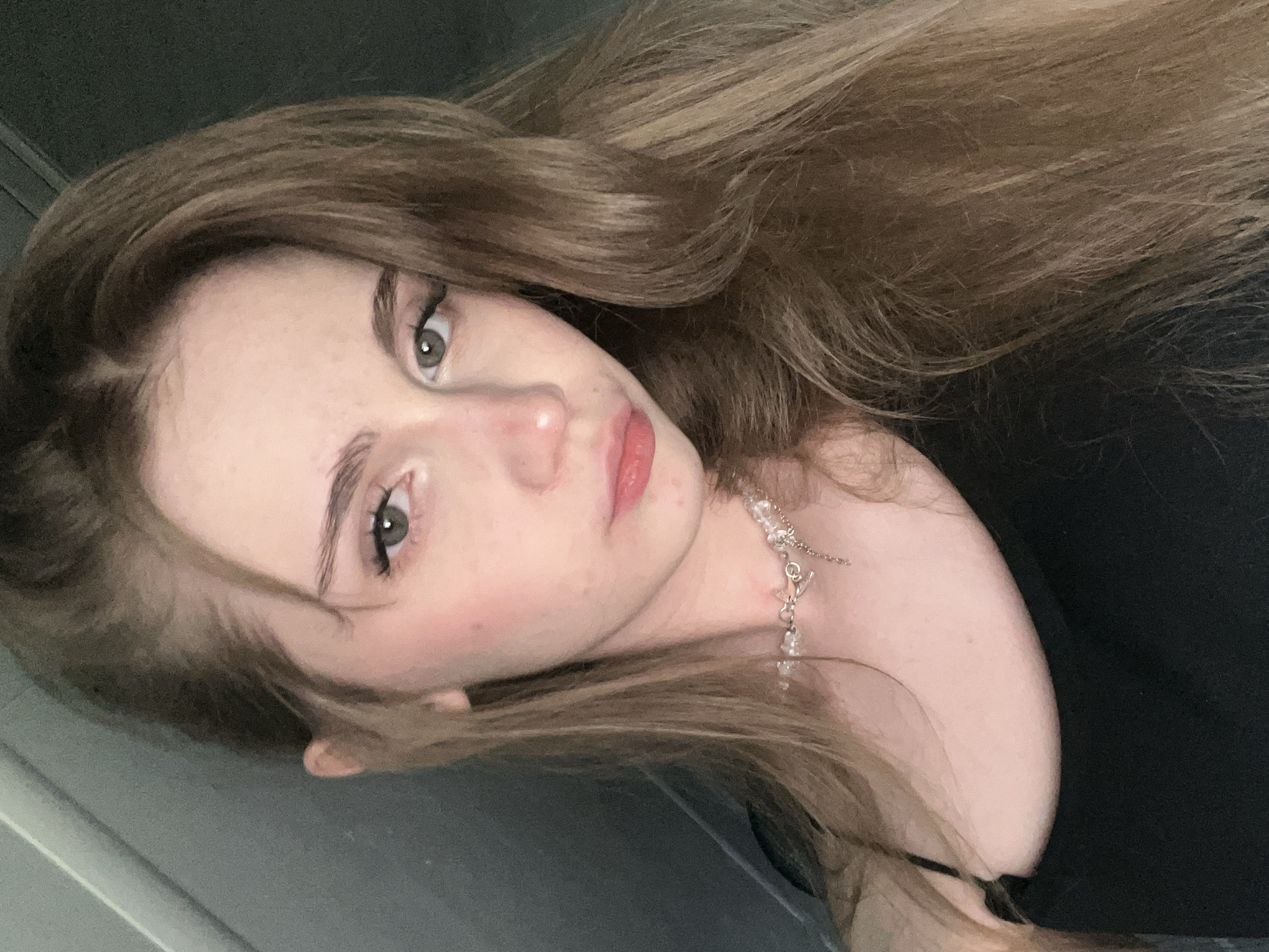 A close-up, rotated image shows a young person with long, light brown hair that is slightly wavy. Their face is tilted towards the camera, with their gaze directed slightly off-camera. They have fair skin with a light dusting of freckles, noticeable rosy blush on their cheeks, and their lips are a soft pink color. Their eyes are a light green-gray, with dark eyeliner accentuating them. A delicate silver necklace with small crystal-like charms is visible around their neck. The background is dark and out of focus, with a hint of gray and dark tones. The lighting appears to be artificial, casting soft shadows on the face. The overall impression is of a casual, introspective moment. No text is visible in the image.