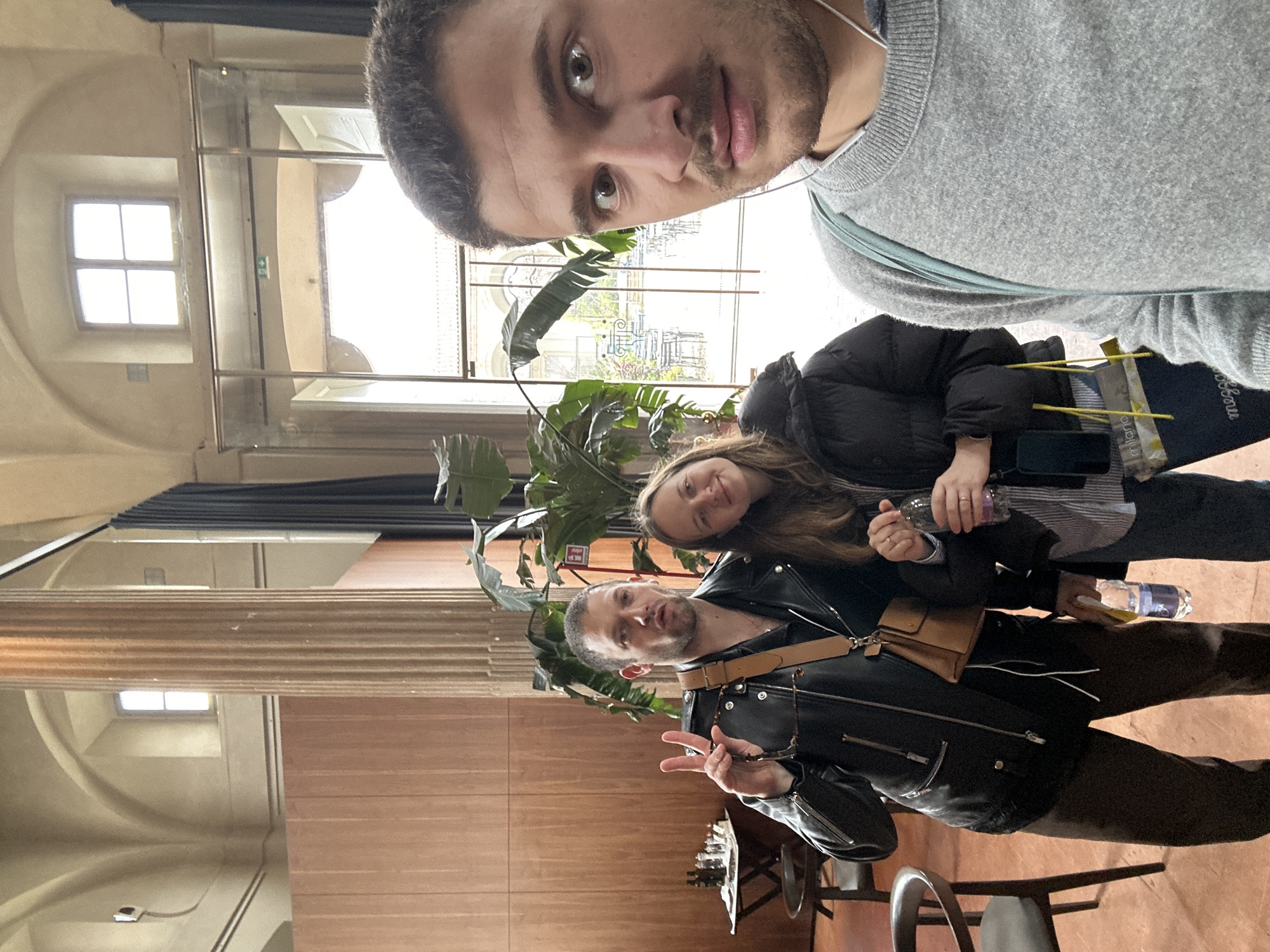 A selfie photograph captures three individuals inside a grand historic building in Milan, Italy, featuring classical architecture and an adjoining outdoor courtyard cafe. The interior displays high vaulted ceilings, light-colored walls, a prominent fluted column, and a reddish-brown floor. Large glass doors in the background open to an outdoor courtyard furnished with numerous round cafe tables and chairs. To the far left, a small round wooden table holds a chessboard and several small bottles, flanked by two wooden chairs. Large green potted plants are arranged between the subjects and the glass doors.
The primary subject, partially visible on the right, is a male with dark hair and stubble, wearing a light gray crew-neck sweater and white in-ear headphones. He takes the photograph, looking directly at the camera with wide eyes and a neutral expression. His head and upper torso occupy the right side of the frame. To his left, in the mid-ground, stands a male with dark hair and stubble, dressed in a black leather jacket over a dark shirt and brown trousers. A tan leather messenger bag with a wide strap is slung across his chest. He holds a pair of sunglasses in his left hand, flashes a peace sign with his right hand, and makes a pursed-lip expression, holding a clear plastic water bottle. Next to him, a female with long brown hair wears a black puffer jacket over a striped shirt. She holds a clear plastic water bottle with a purple cap in her right hand and a dark blue shopping bag with yellow strings, partially displaying "milano" and "mezzogiorno," with a smartphone tucked into her left jacket pocket. She smiles faintly, leaning slightly towards the male figure on her left.