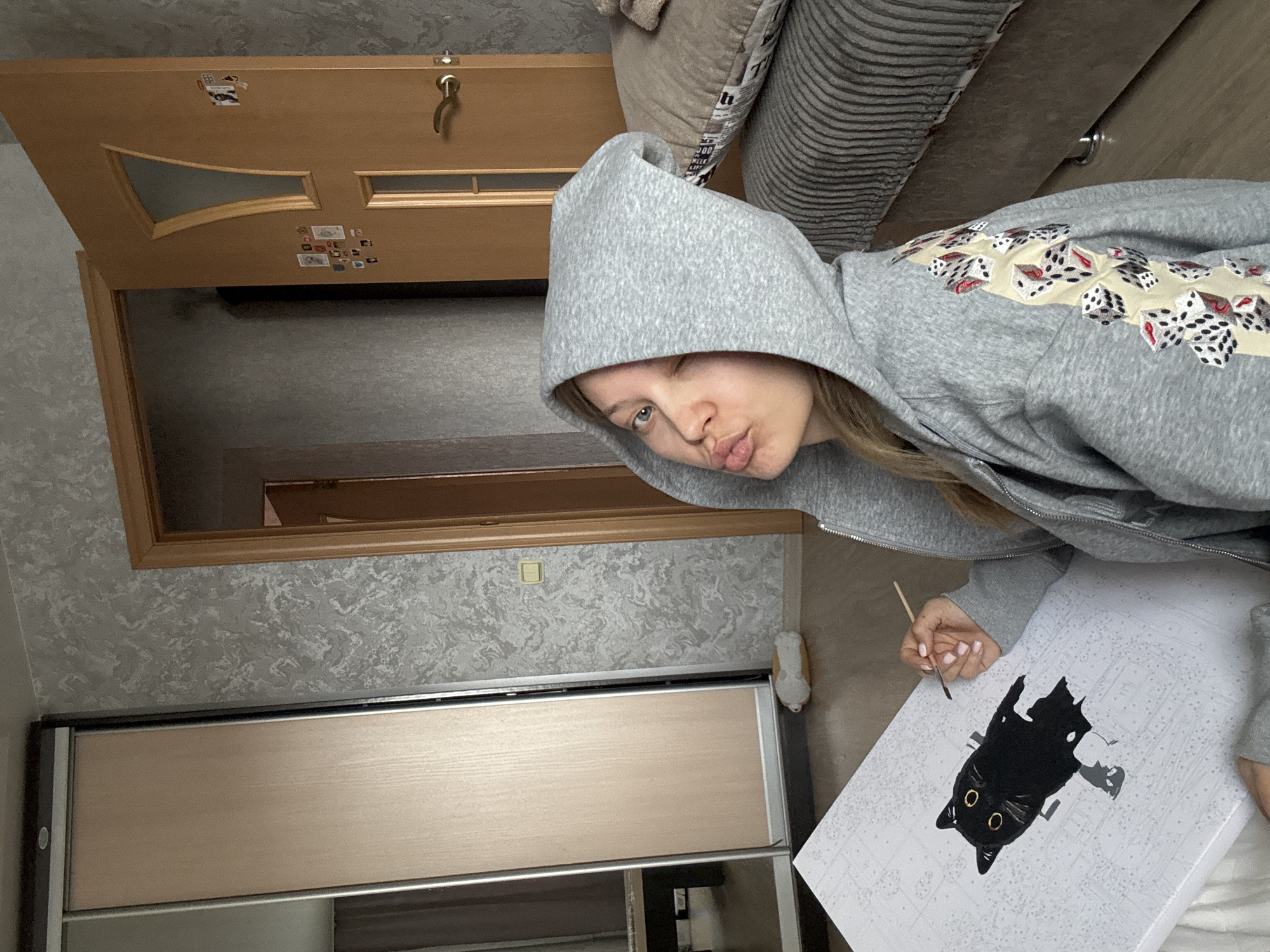 The image features a young person, likely female, engaged in painting by numbers within a residential setting in Chaikovsky, Russia. She is wearing a grey hooded sweatshirt with the hood pulled up, and her hair is visible. Her face is turned towards the camera, winking with one eye and pouting her lips in a playful expression.
In her hands, she holds a small paintbrush, actively painting on a canvas that depicts a partially completed black cat with bright yellow eyes. The cat image on the canvas is oriented upside down relative to the viewer. The grey sweatshirt is notable for a distinctive decorative band across the shoulder, featuring a repeating pattern of red and black dice on a lighter-colored strip.
The background reveals parts of a room. To the left, a light brown wooden door with glass inserts is slightly ajar, adorned with several small stickers or photographs. The wall behind the door and above a wardrobe is covered in light grey wallpaper with a subtle, mottled pattern. Below this, a section of a sliding wardrobe or cabinet with light wooden panels and a dark frame is visible. In the upper right corner, a portion of a bed or couch with textured, patterned covers can be seen, where faint text like "WEEK" and "MILK LIFE" is discernible. An electrical outlet or light switch is also visible on the patterned wall. The scene captures a relaxed, creative moment indoors.