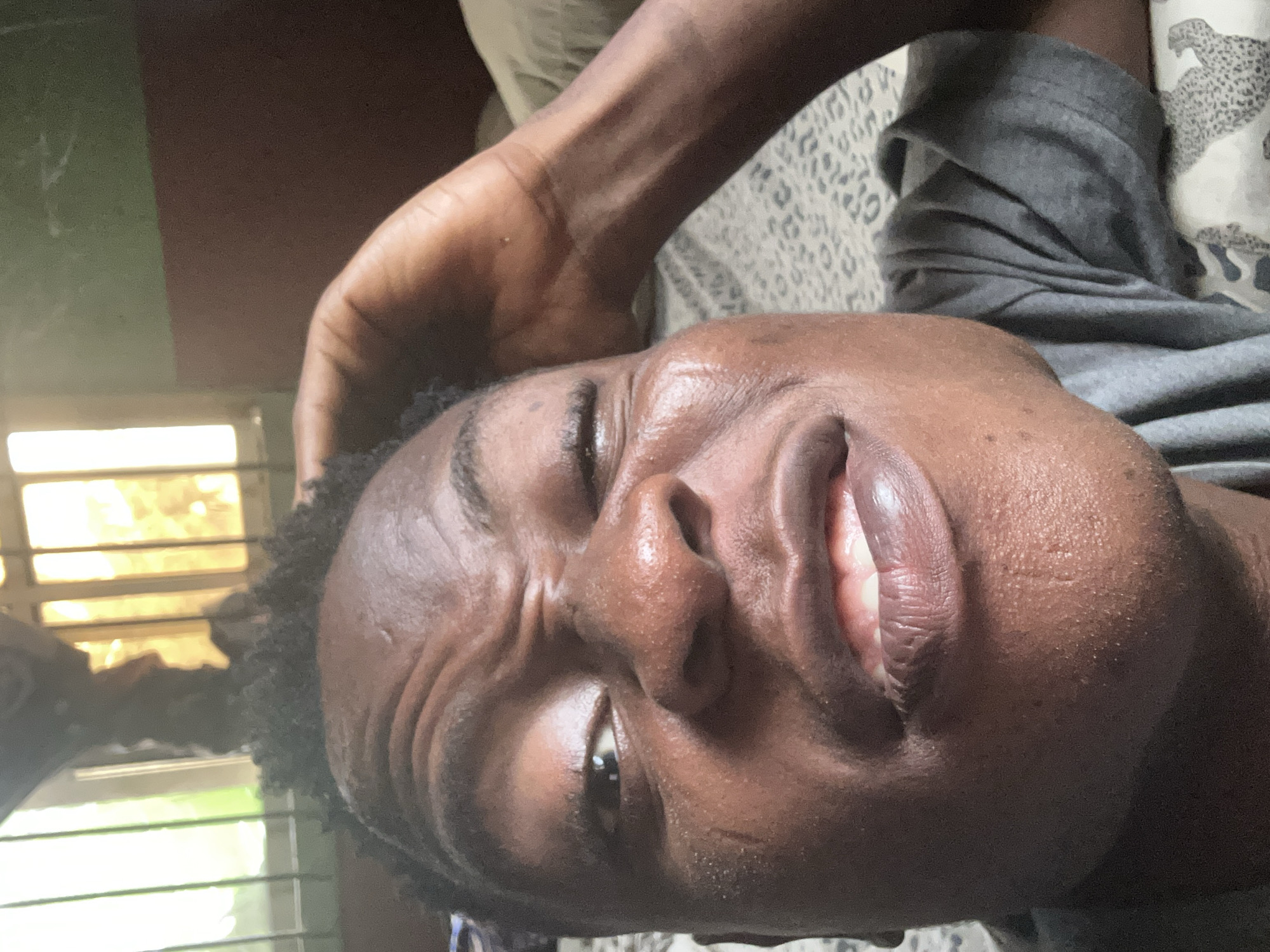 A close-up, angled selfie shows a person with dark skin and dark hair styled in short dreads. Their face is contorted into an exaggerated expression, with the right eye squinted shut and the left eye open, looking towards the camera. Wrinkles are prominent on their forehead and around their eyes, suggesting the forceful facial expression. Their mouth is slightly open, revealing teeth and a tongue. The person's right hand is raised and resting on their head, with fingers splayed in their hair. They are wearing a dark grey t-shirt. The background includes a bed with patterned sheets and a pillow. To the left, a window with multiple panes is visible, with sunlight filtering through, creating a bright, hazy effect. A two-toned wall, with a green upper section and a reddish-brown lower section, is also present in the background. The scene appears to be indoors, possibly in a bedroom, given the presence of a bed and window. The overall impression is of a casual, self-taken photograph.