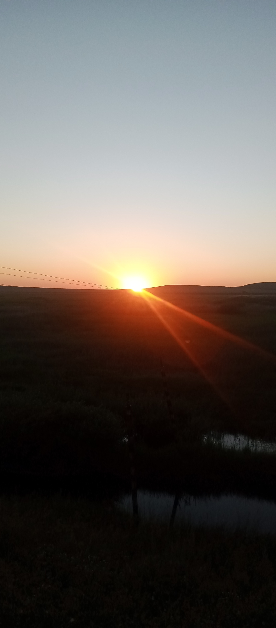 A vertical photograph captures a sunset over a rural landscape in Marinovka, Kazakhstan. The sun, a bright orb, is just beginning to dip below the horizon, casting a warm, orange glow across the sky. Sunbeams radiate outwards from the sun, creating a lens flare effect. In the foreground, a dark, grassy field stretches out, with a small body of water, likely a pond or ditch, visible in the lower portion of the frame. A few fence posts are sparsely scattered throughout the field, adding to the rustic feel of the scene. The sky transitions from a light blue at the top to a soft orange near the horizon, indicating the late hour of the day. The overall mood is serene and peaceful, conveying the quiet beauty of a country sunset. There is no visible text in the image.
