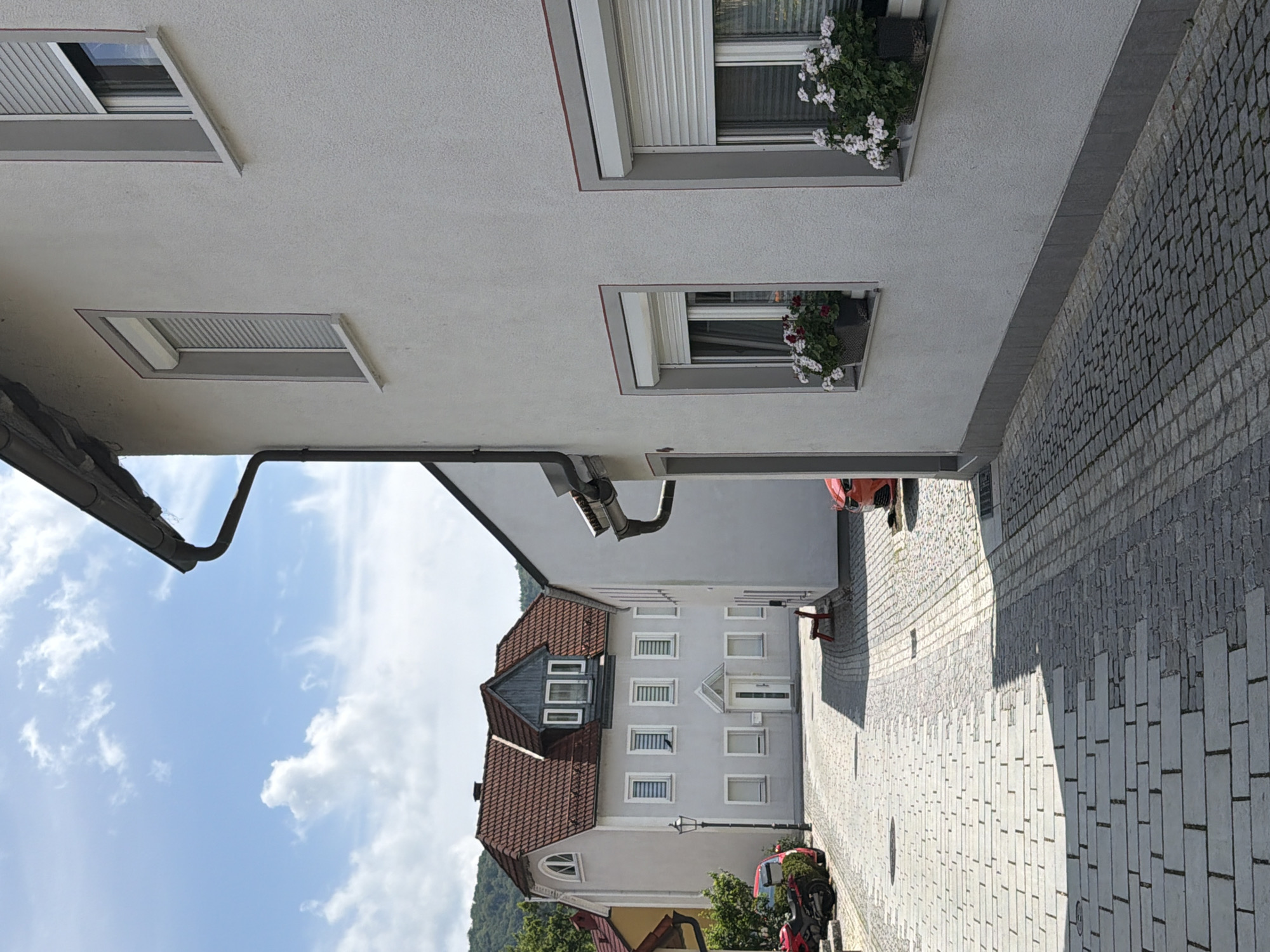 The image shows a street scene in Ebermannstadt, Germany, with a cobblestone road leading towards a building with a red tiled roof. The building on the right is white with a textured facade and has two windows with flower boxes. The street is paved with interlocking cobblestones and has some shadows cast by the buildings. In the background, there is another white building with multiple windows. A red motorcycle and a red car are parked on the left side of the street. The sky is blue with scattered clouds.