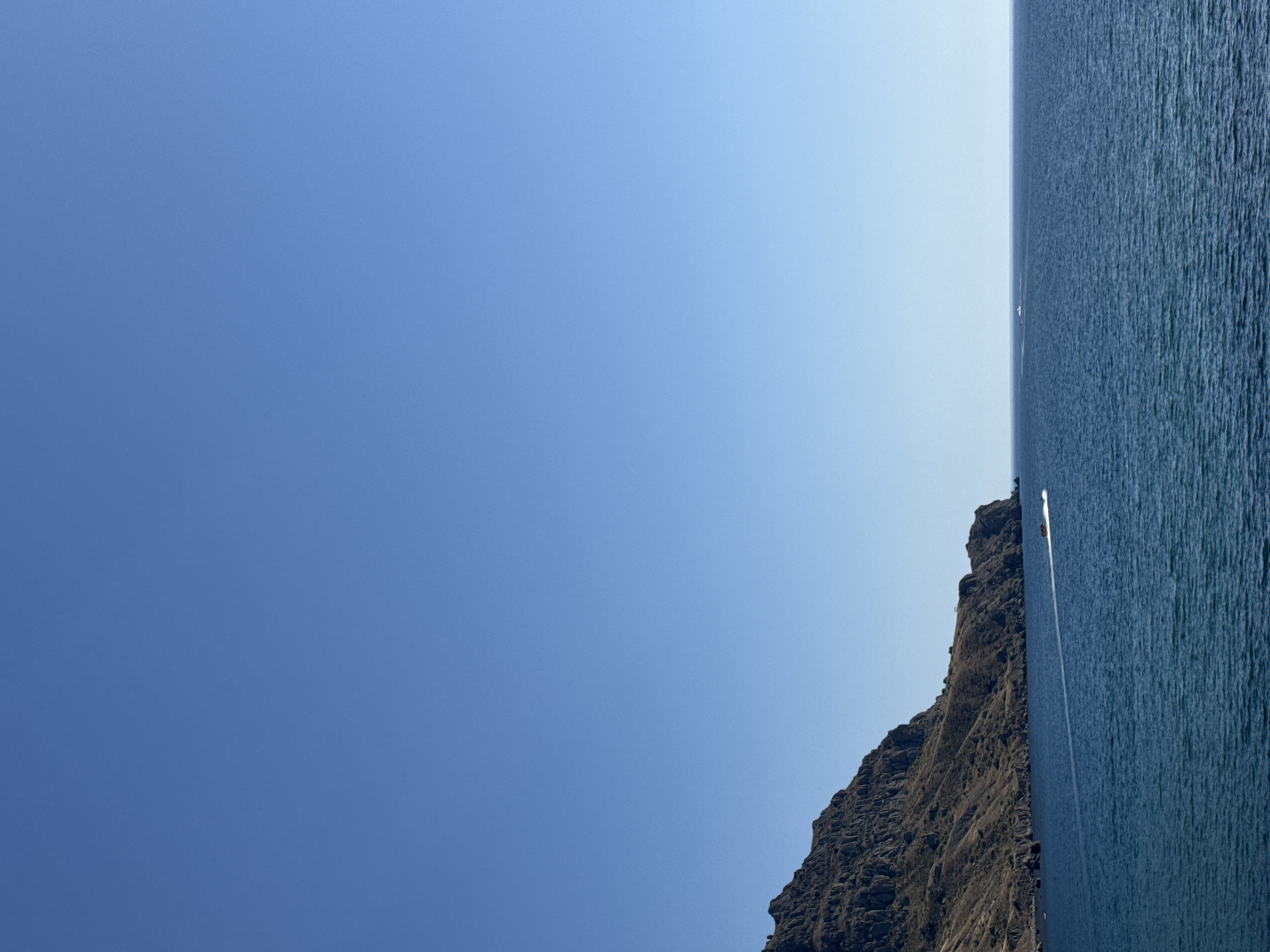 The image shows a vast expanse of calm, deep blue ocean under a clear, cloudless sky. To the left, a rugged, dark brown mountain range extends into the water, with scrubby vegetation clinging to its slopes. A white wake trails across the ocean, indicating the path of a small, fast-moving boat with at least two visible occupants. The boat is positioned in the lower middle section of the image, creating a dynamic element against the stillness of the sea and sky. The horizon line is sharp, separating the deep blue of the water from the pale blue of the sky. No specific urban features are visible, suggesting a natural coastal landscape.