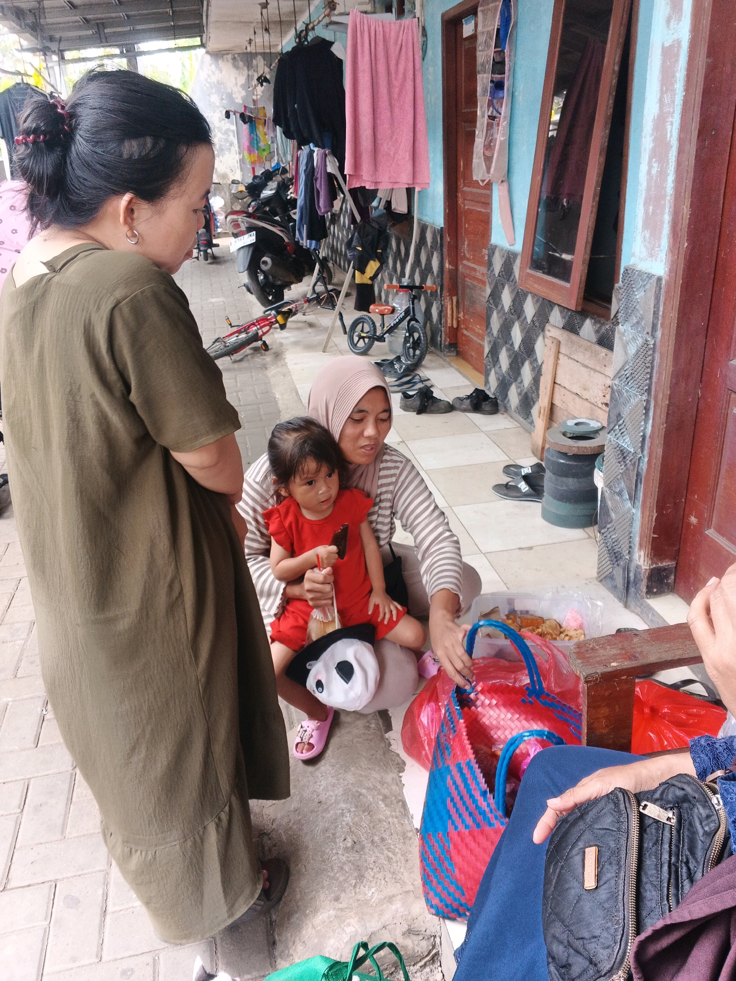 An adult female with dark hair pulled back in a bun stands to the left, wearing a long, olive-green dress. She is looking down and to her right. Seated on the ground in the center is another adult female, wearing a light-colored, striped long-sleeved shirt and a light pink hijab. She is holding a young child on her lap, who is wearing a red outfit and pink sandals. The child is holding a dark item, possibly food. Next to the seated adult, a woven red and blue bag is open, and a clear plastic bag contains various food items. To the right of the seated adult, another person is partially visible, wearing a blue garment and holding a black quilted pouch. The scene appears to be outdoors in front of a building with blue and light blue painted walls, a wooden door, and a window with a dark frame. The ground is paved with light-colored square tiles. In the background, motorcycles and clothing hanging on a rack are visible, suggesting a street or market area in Kadu, Indonesia.