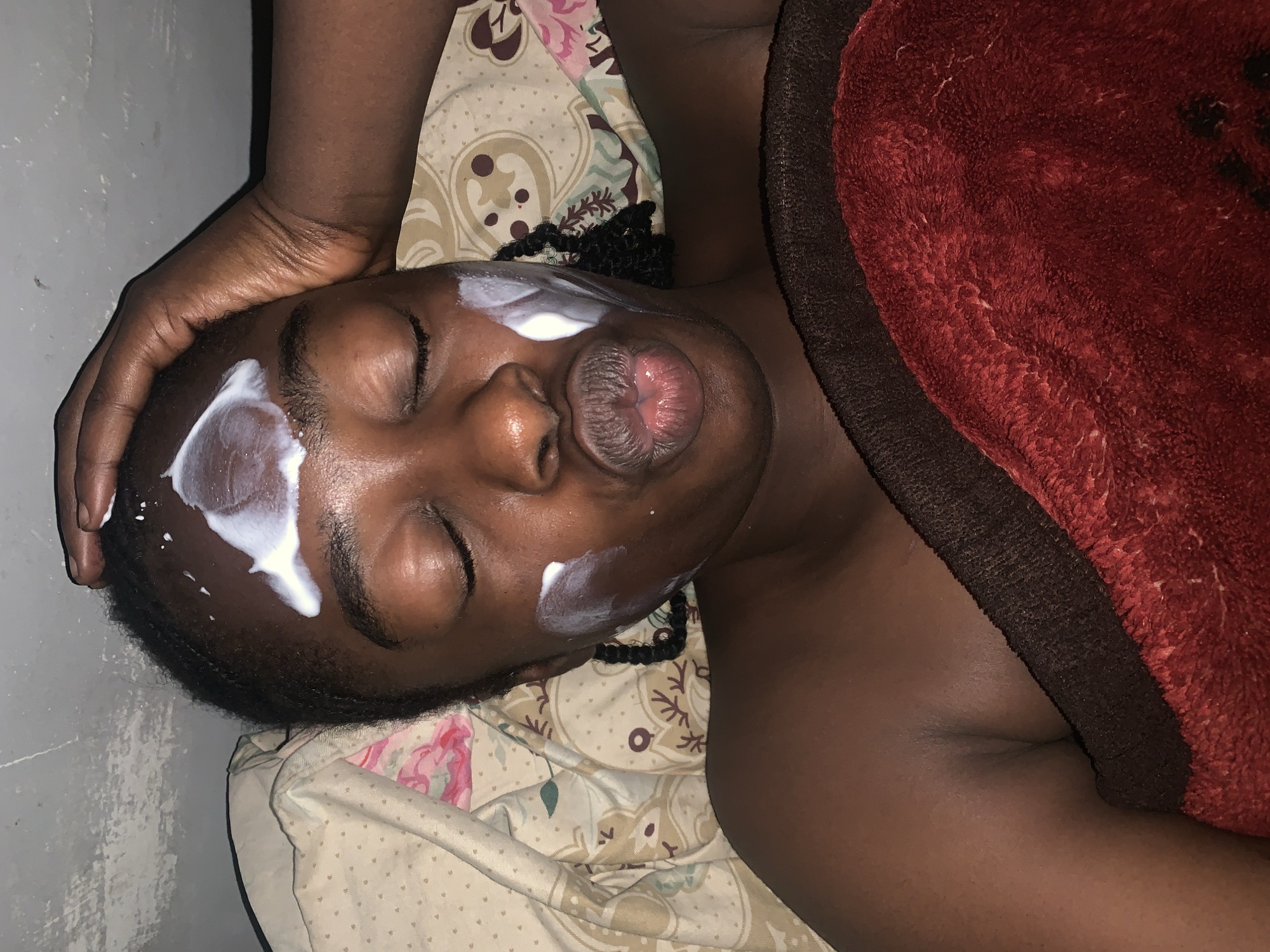 A dark-skinned individual lies supine with eyes closed and lips pursed into an O-shape, indicating relaxation or a specific facial exercise. White cream is applied in distinct streaks and dots on their forehead and right cheek. A dark-skinned hand, likely belonging to another person, gently rests with fingers extended on the individual's forehead and temple. The individual has short, dark, textured hair, with some braided elements visible near the neck area. They are resting on a patterned bedsheet featuring beige fabric with a repeating motif of pink flowers, green leaves, and dark brown circular patterns. A deep red, fuzzy blanket covers a portion of their body from the chest downwards, with a dark brown, textured fabric visible underneath the red blanket. To the left of the individual's head, a textured light grey surface, possibly a painted wall or concrete floor, is visible. The scene is located in Rafin Par, Nigeria.