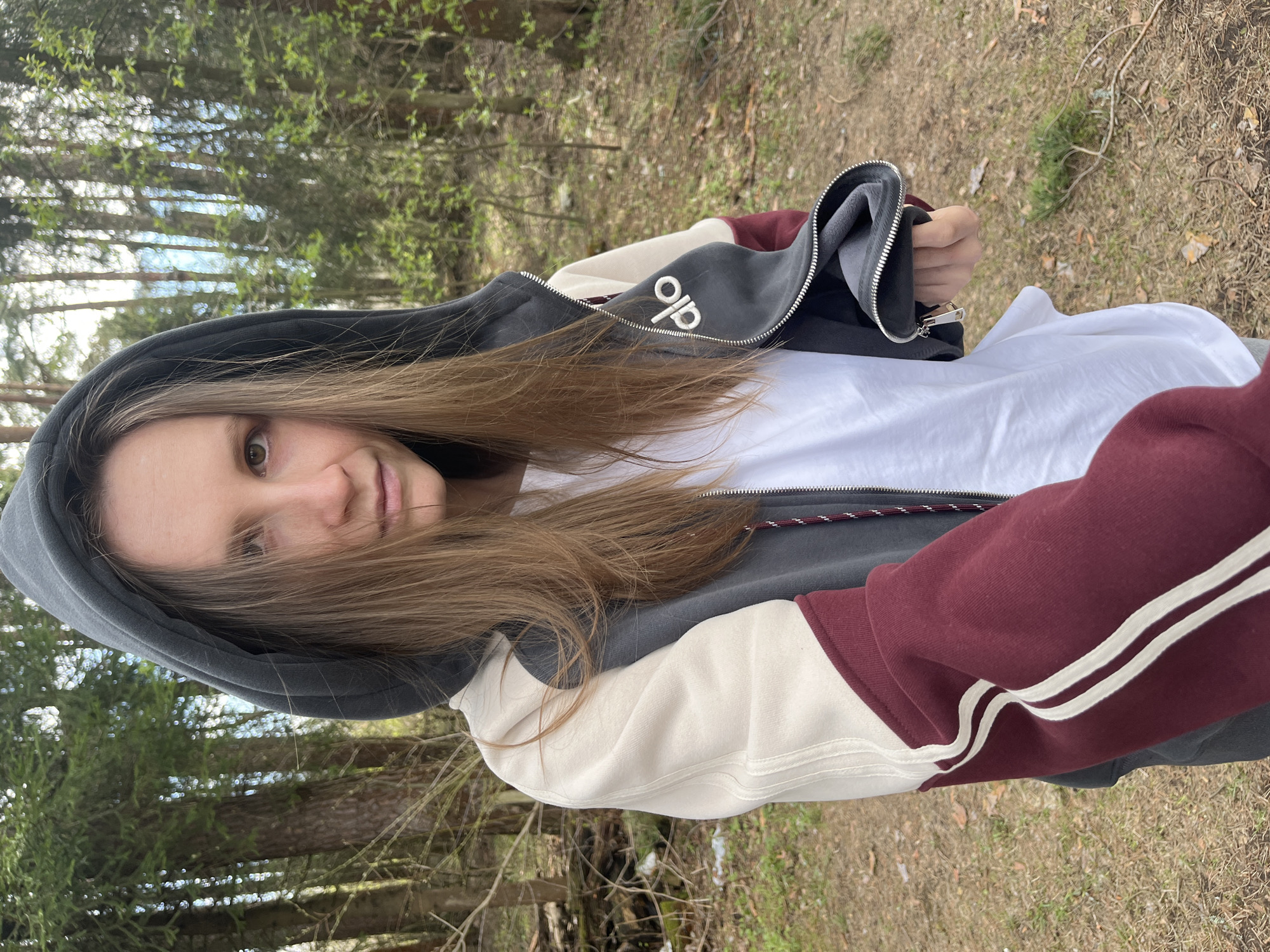 A young woman stands outside, possibly in a wooded area near Раздолье, Russia, as suggested by the pine trees visible in the background. She is wearing a hooded zip-up jacket with color-blocked sleeves and a white T-shirt. The jacket has the word "alo" embroidered on the chest. Her hair is brown and falls past her shoulders, obscuring part of her face. The ground is covered with dry grass and fallen leaves.