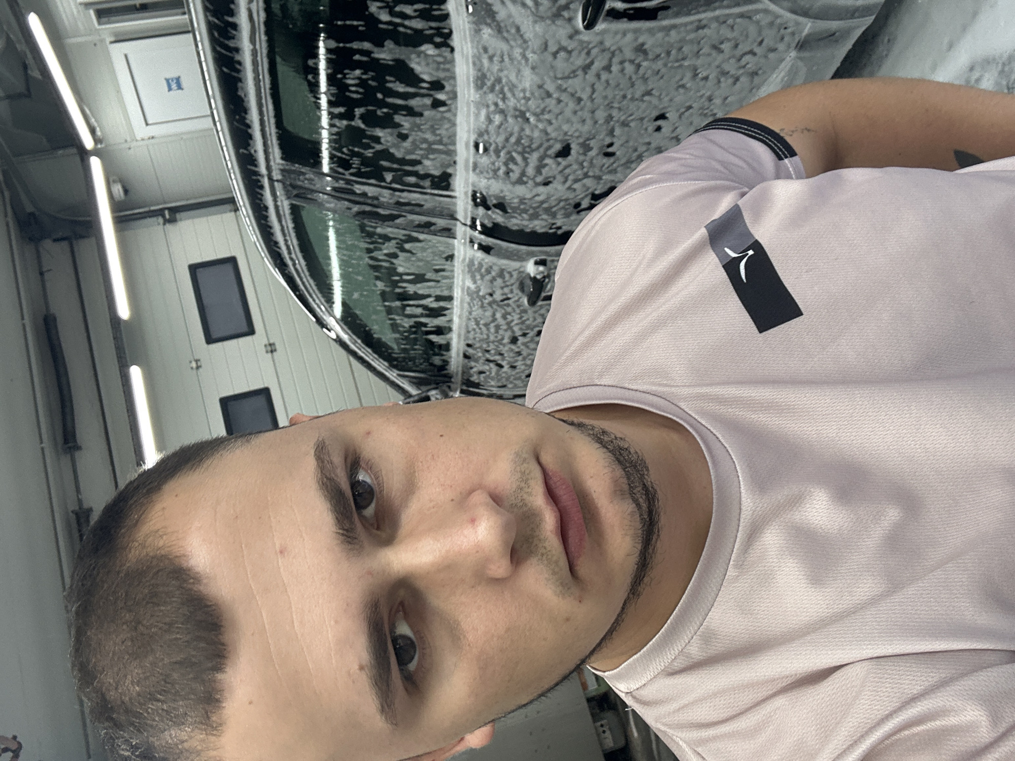 A close-up, selfie-style photograph features a man with short, dark hair, a light beard, and a serious expression. He is wearing a light pink t-shirt with a black and gray rectangular emblem on the left breast. To the right of the man, a dark-colored car is partially visible, covered in white foam, suggesting it is being washed. The background appears to be a garage or car wash facility, with bright fluorescent lighting and industrial-style doors. No specific location in Chișinău, Moldova is identifiable from the image itself.
