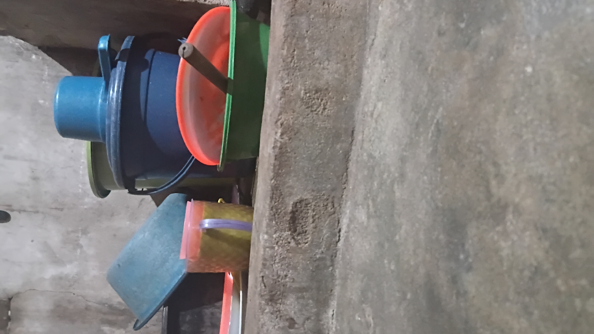 In a dimly lit interior, likely a storage area in Prabumulih, Indonesia, a collection of plastic containers and buckets are arranged on a concrete ledge and the floor. A large, dark blue bucket with a blue lid is prominent, partially obscuring a lighter green bucket behind it. To the left, a turquoise basin is tilted, resting on an unseen surface. In front of the buckets, a yellow and clear plastic container with a purple handle is visible. Further to the right, a green basin holds an orange lid, with a brown stick resting on it. The background consists of a textured, pale gray wall, showing some signs of wear and cracking. The foreground is dominated by a rough, textured concrete floor and ledge. The lighting suggests artificial illumination, casting subtle shadows. No individuals are present in the scene.