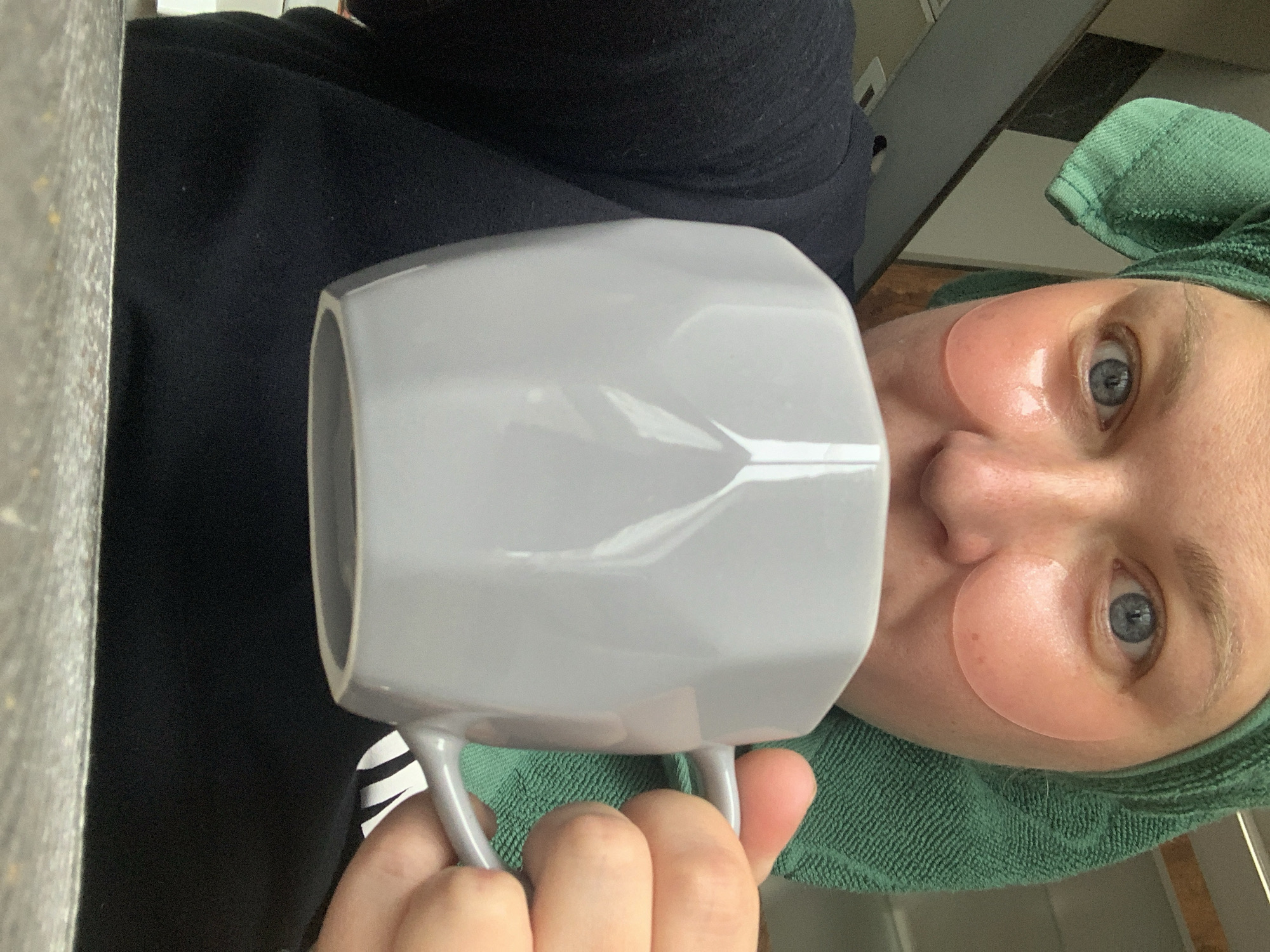 A person with fair skin is holding a light gray polygonal mug up to their face. The person has a green towel wrapped around their head and peach-colored under-eye masks applied. They are wearing a dark-colored shirt. A gray surface is visible to the left. The photo appears to be taken indoors. There is no information provided that can indicate that this image was taken in the city of Chaikovsky, Russia.