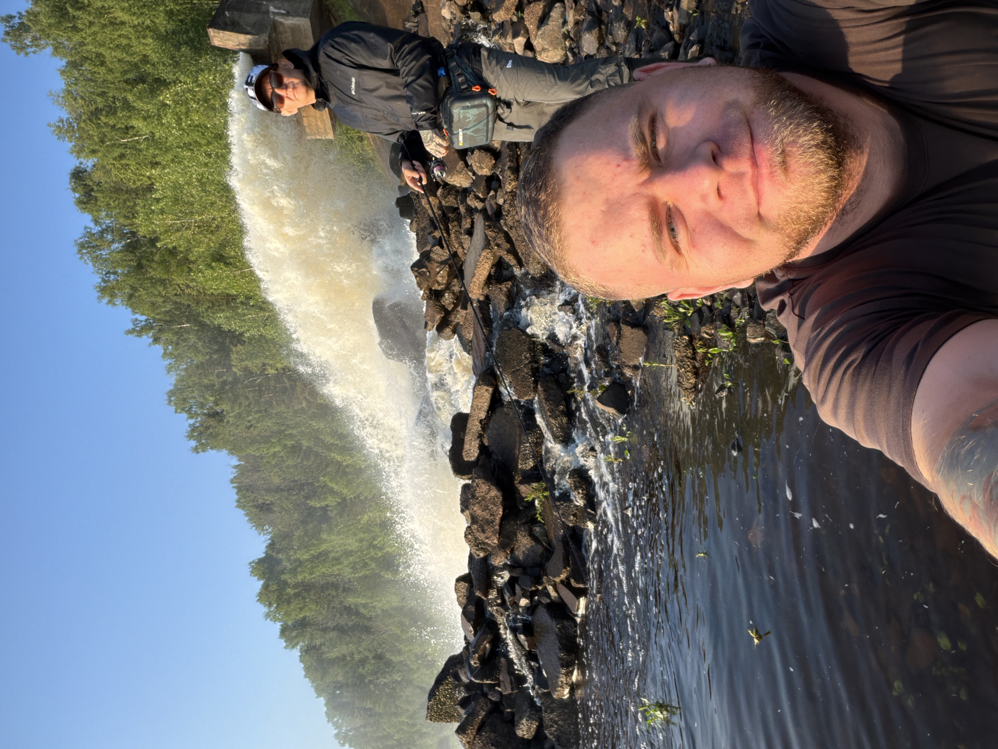 The image shows two men near a powerful water discharge into a rocky stream. The man in the foreground is taking a selfie, showing his face and upper body. He has light skin, a beard, and is wearing a brown t-shirt. The man in the background is standing to the right, facing left and looking towards the camera. He is wearing a dark jacket with a "Columbia" logo, grey pants, a cap, and sunglasses. He appears to be fishing, holding a fishing rod with a reel. A green and black pouch is attached to his waist. Behind them, a large volume of water is being discharged, creating a spray and turbulent flow. The water is flowing over and around large rocks that form a barrier in the stream. Dense green trees line the background, suggesting a forested area. The sky is clear and blue. This scene is located at the Chernushka Hydroelectric Power Station in Chernushka, Russia, as evidenced by the unique concrete structure and the powerful water outflow. The lighting suggests it is daytime, likely morning or late afternoon due to the warm sunlight.
