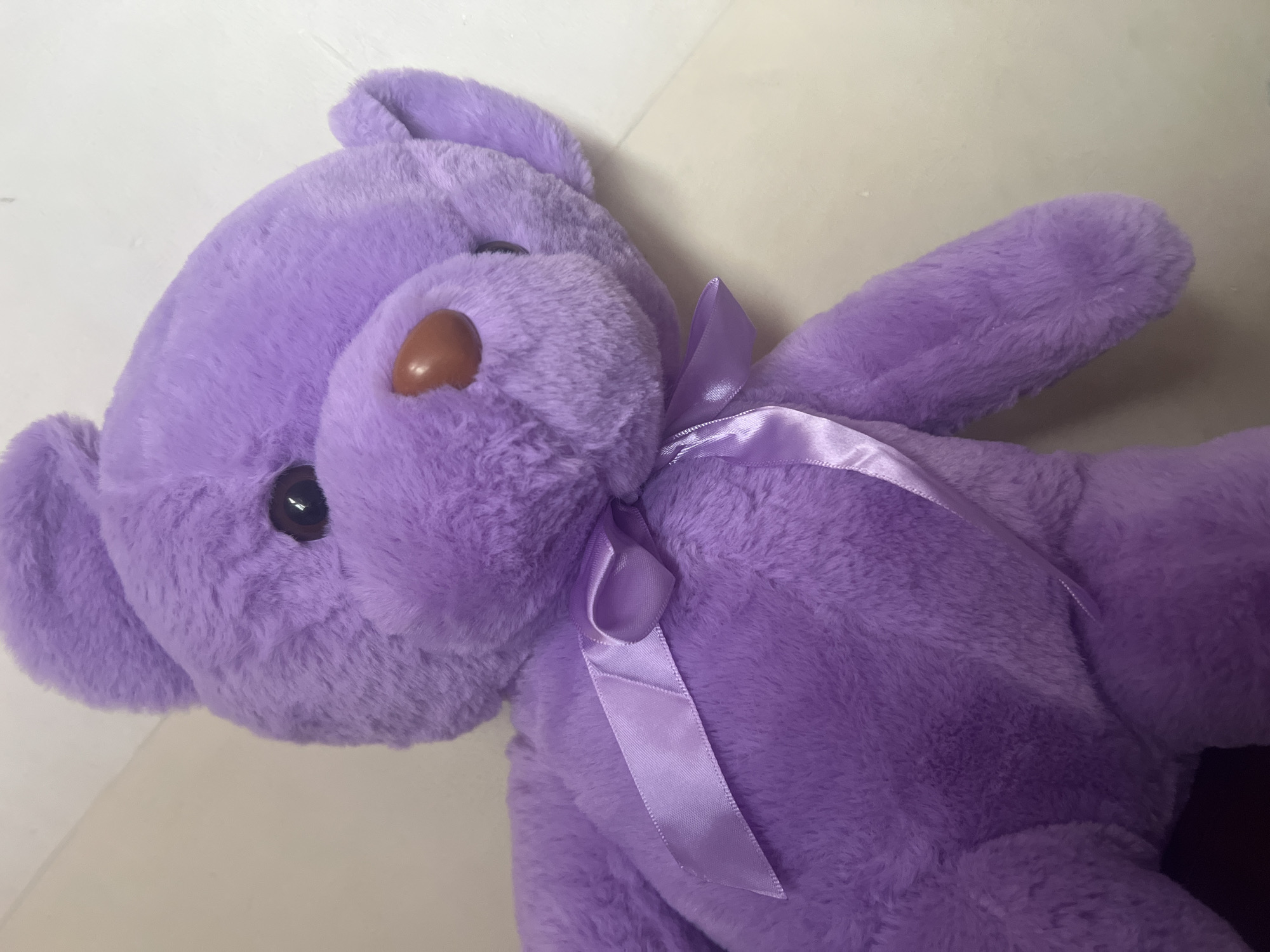 A plush, purple teddy bear with a brown nose and a satin bow around its neck lies on a flat, light-colored surface. The bear is positioned at an angle, with its head and left ear prominently displayed.
There is no evidence to suggest this image was taken in the city of Dar es-Salaam, Tanzania.