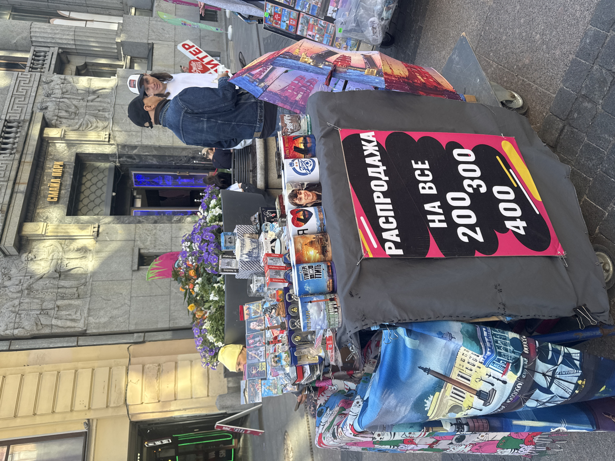 A street vendor displays merchandise on a cart in front of a building in Saint Petersburg, Russia. The building has a facade with classical architectural elements, including columns and a relief sculpture above an entrance. Above the entrance, a sign reads "СМАЙЛ ПАРК" in Russian.
The vendor's cart is laden with various items for sale, including postcards, magnets, mugs, and decorative objects. A bright pink and black sign on the cart advertises a sale with prices listed as 200, 300, and 400, and text that translates to "SALE ON EVERYTHING." Colorful textiles, possibly scarves or fabric hangings, are draped over the front of the cart. In the foreground, there are cobblestone pavers.
Several people are visible in the scene. Two men are standing near the cart, and other individuals are seen in the background, some sitting and others walking. One of the men near the cart is wearing a denim jacket and a black baseball cap. The other man is wearing a white t-shirt and a baseball cap. A partially visible person in the background is wearing a yellow beanie. The overall impression is a bustling street scene with a vendor selling souvenirs and gifts.