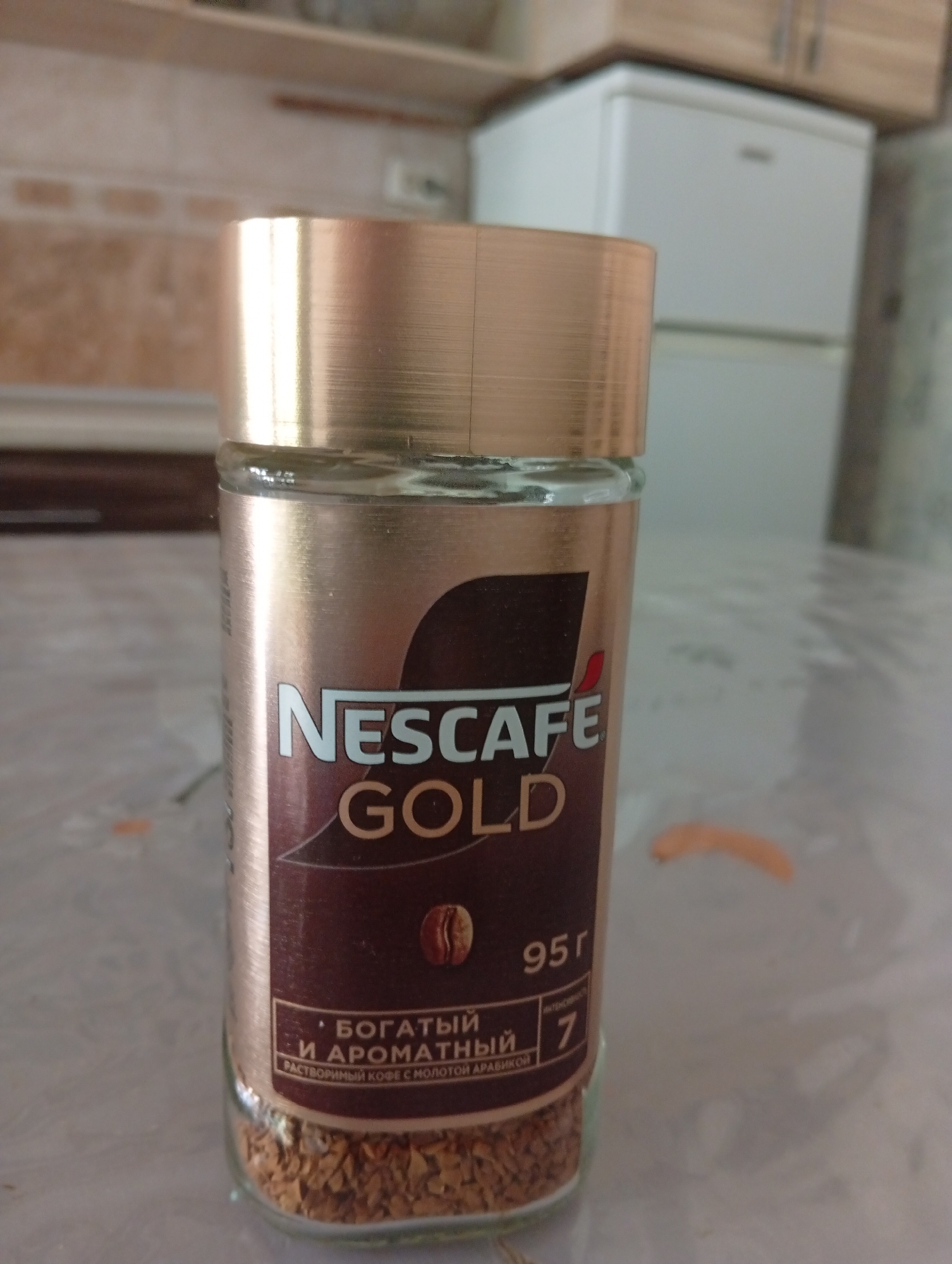 A close-up shot focuses on a jar of Nescafé Gold coffee. The jar, made of glass with a metallic gold lid, is filled with instant coffee granules. The label on the jar is predominantly gold and brown, displaying the Nescafé Gold logo and the text "БОГАТЫЙ И АРОМАТНЫЙ" (Rich and Aromatic) in Russian, along with "95 г" (95g) indicating the net weight. The background shows a blurred kitchen environment with cabinets and a refrigerator. The scene is indoors, on a tabletop, and the lighting appears to be natural, possibly from a window. There are no people visible in the image. The image does not provide any specific details about the city of Chigatay, Uzbekistan, or its features.
