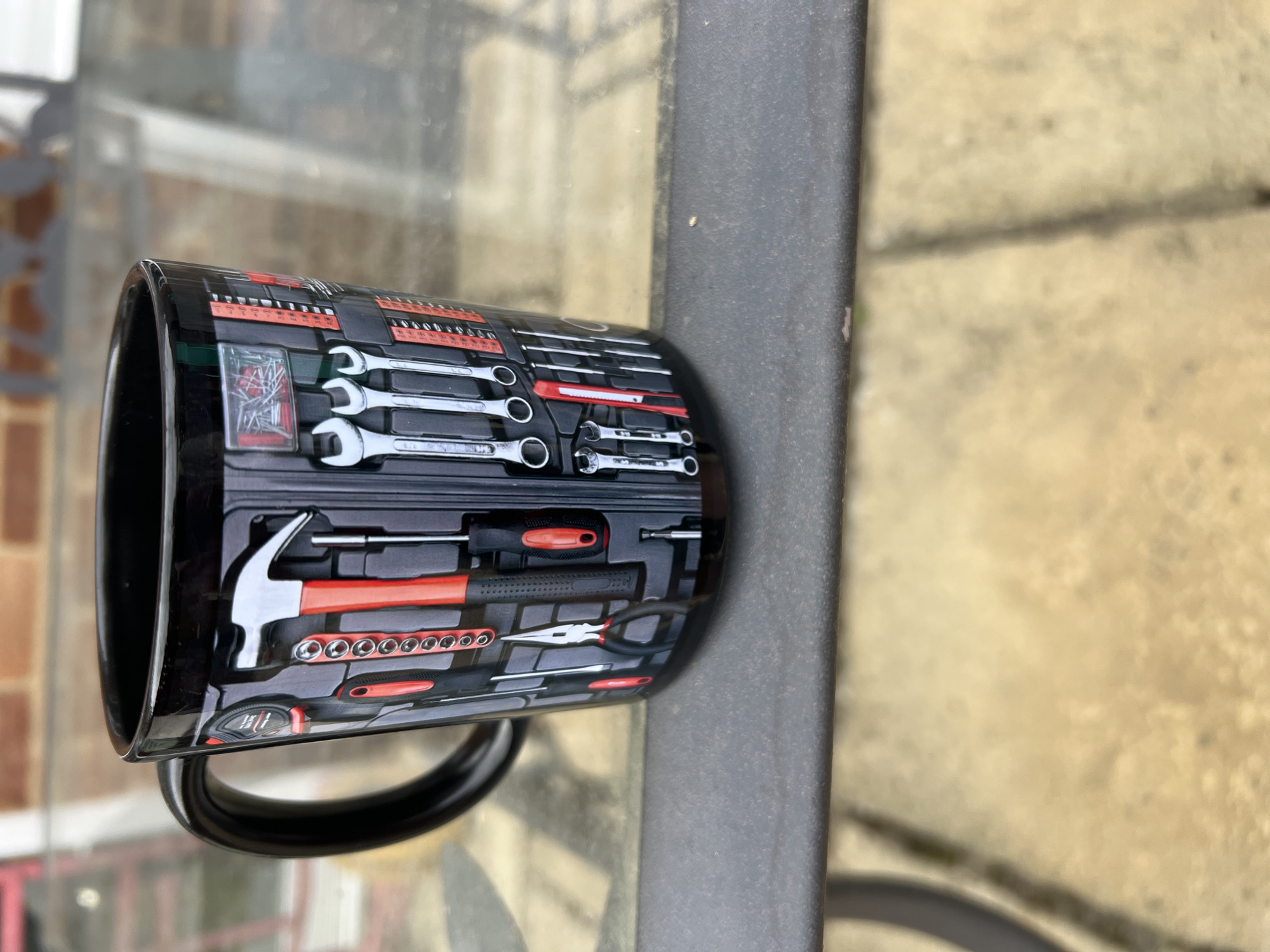 A black mug with a tool-themed design sits on a gray surface, likely a metal table, outdoors. The mug's design features an array of tools including wrenches, screwdrivers, a hammer, and various other implements arranged as if in a toolbox. The background shows a partially blurred view of a building with brick details and a glass surface reflecting outdoor elements. The scene is outdoors and the location cannot be determined with any certainty.