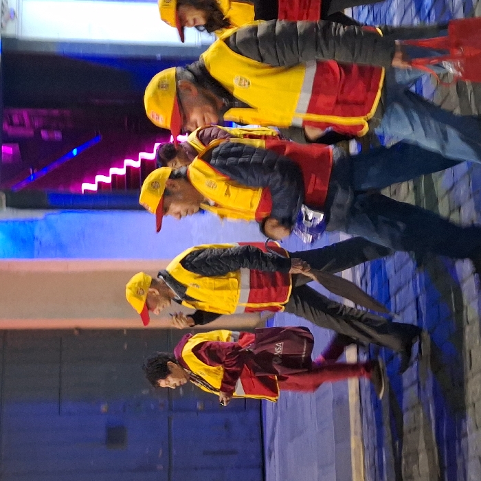 A line of people wearing yellow hats and vests are walking. The vests have red trim, and some have a red backing. A person at the bottom of the frame is wearing a maroon outfit with yellow accents. Behind the group, there's a wall with blue panels and bright pink stair-like lights above. They appear to be walking along an outdoor walkway or street.