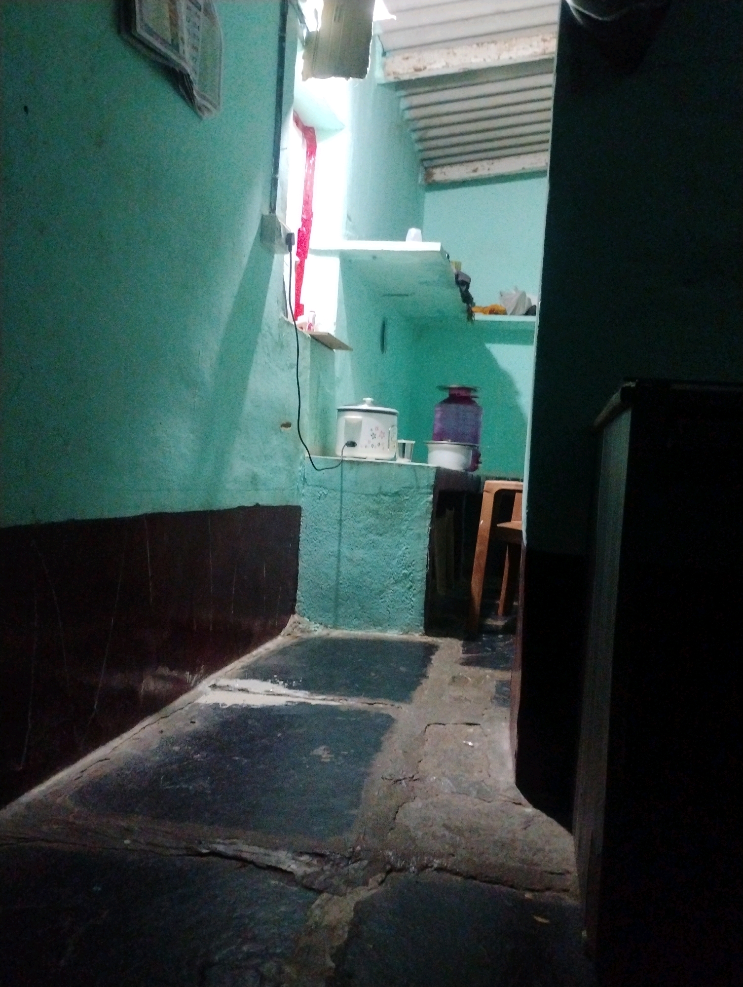 The image depicts a narrow, dimly lit interior passageway, likely in a residential setting within Bodana Hosahalli, India. The walls are painted a light teal color. On the left, a dark brown wainscoting runs along the lower portion of the wall. The floor appears to be made of large, irregularly shaped stone slabs, some of which are cracked and worn.
Towards the back of the passageway, on a textured teal ledge or shelf, sits a white rice cooker with a patterned design. Adjacent to it is a purple water jug with a white lid. A small wooden stool with a reddish-brown finish is positioned to the right of the ledge. Further back, a sliver of natural light enters from a window or opening, illuminating a section of the teal wall. The upper part of the passageway features a corrugated metal ceiling. On the far right of the image, a dark, unidentifiable object, possibly furniture or a doorway, is visible. A black cable hangs down from the ceiling area on the left. A framed document or calendar is affixed to the teal wall on the upper left.
