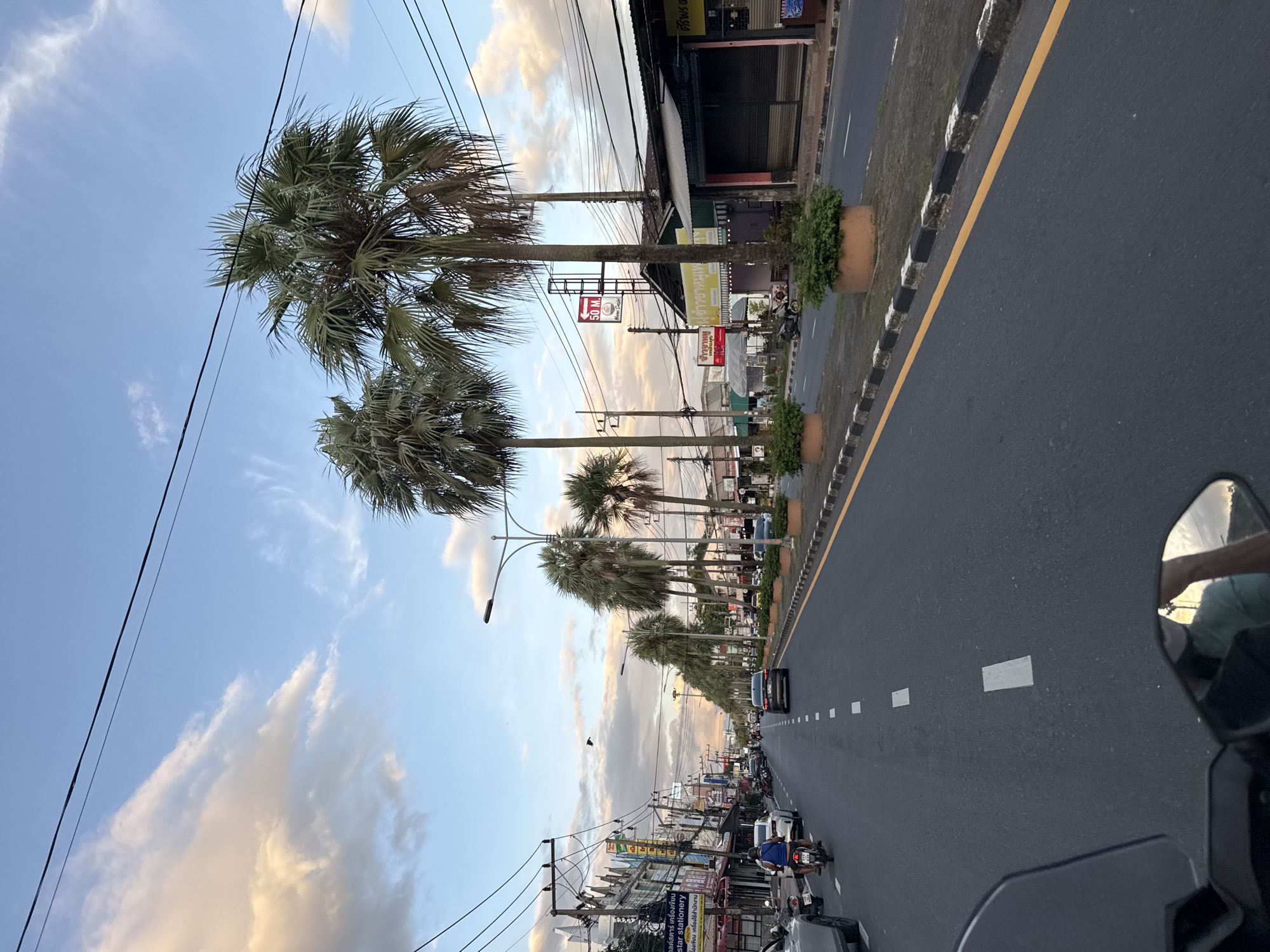 The image depicts a street scene in Wichit, Thailand, during the golden hour, likely late afternoon or early evening, as indicated by the soft, warm lighting and the colors in the sky. The perspective is from a vehicle, possibly a motorcycle, looking forward down a two-lane road. Palm trees line the right side of the street, interspersed with planters containing greenery. Buildings, many with storefronts and signs in Thai script, occupy the left side of the road. Cars and motorcycles are present on the road, with one motorcycle rider prominently visible in the foreground. Overhead power lines crisscross the sky. The sky itself is a mix of blue with wispy clouds tinged with orange and yellow from the setting sun. The road surface is asphalt with white dashed lane markings and a yellow line along the right edge.