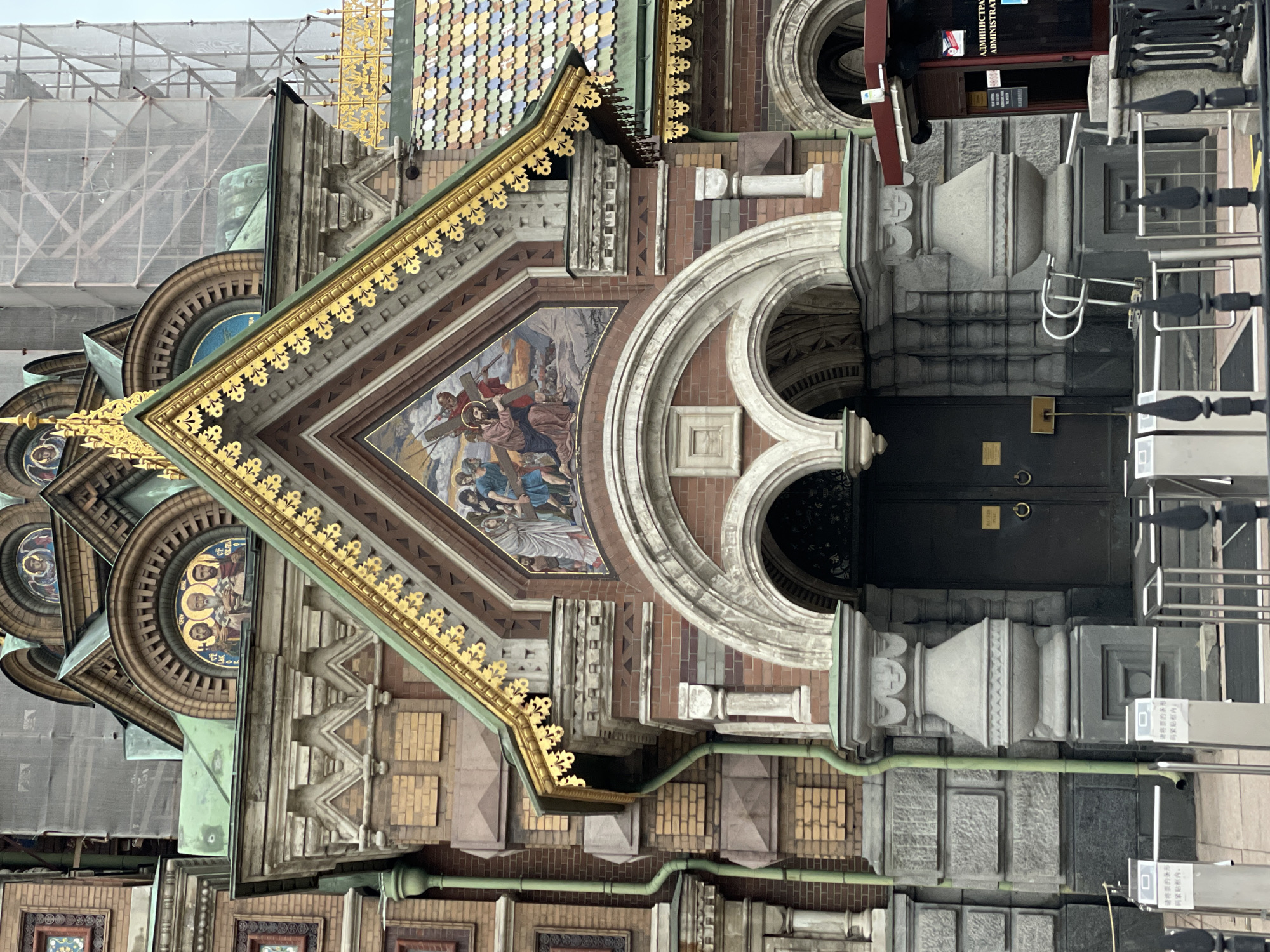 The photograph features an exterior view of the Church of the Savior on Spilled Blood in St. Petersburg, Russia. The focus is on a portion of the building's facade, showcasing the intricate architectural details, including a mosaic depicting a biblical scene under a triangular gable. The building exhibits a mix of brickwork, stonework, and decorative elements, such as arched windows and gold trim. A set of black doors and nearby turnstiles suggest an entrance point for visitors. The presence of scaffolding and protective material hints at ongoing restoration work. Signage in both Russian and another language (possibly Chinese) is visible, indicating the building's administrative or operational details. The overall composition is captured from a relatively close perspective, highlighting the textures and patterns of the building's materials.