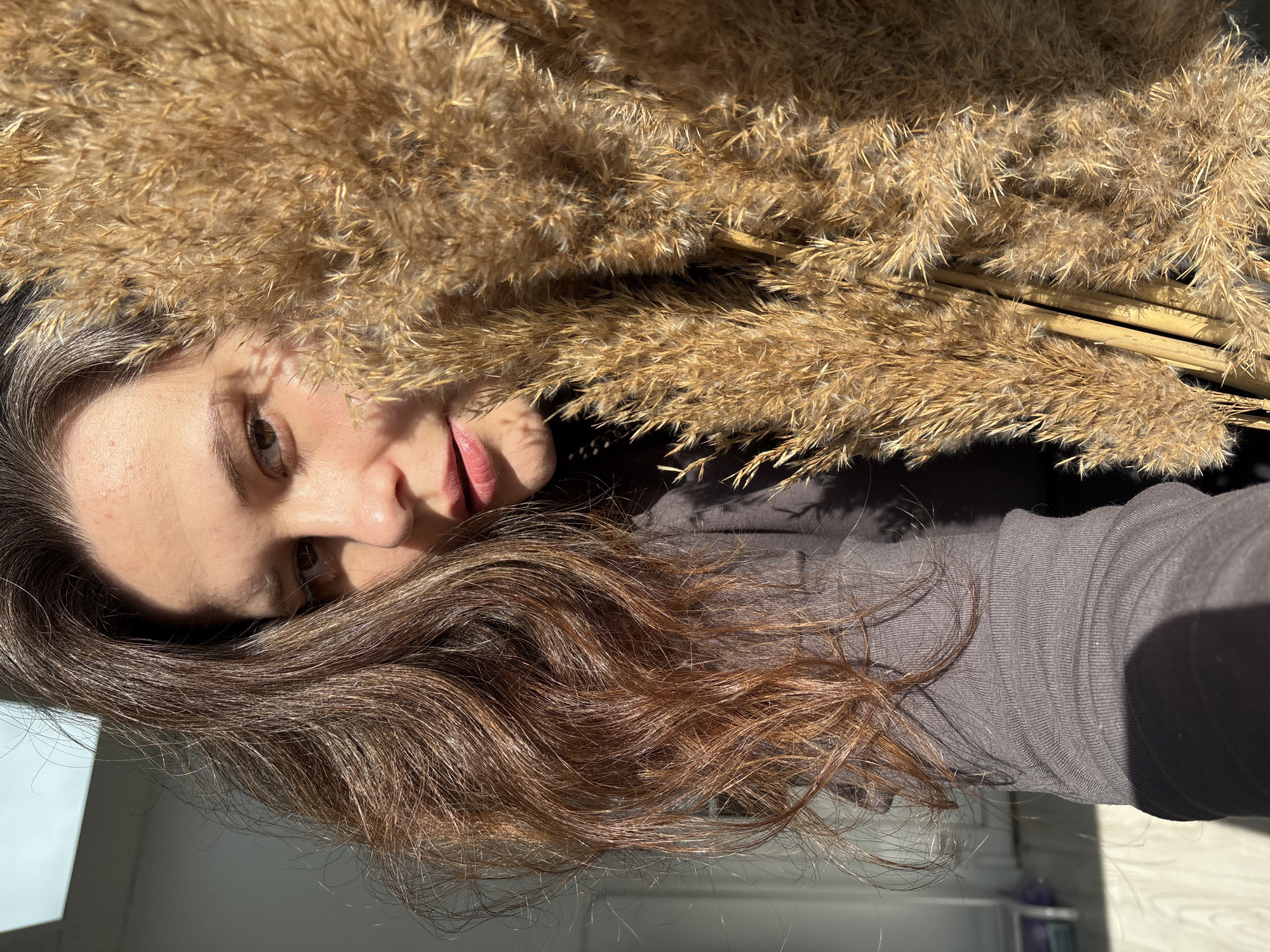 A woman with brown hair lies next to a bundle of pampas grass. The location is likely indoors, possibly in a home. The city is Chaikovsky, Russia.