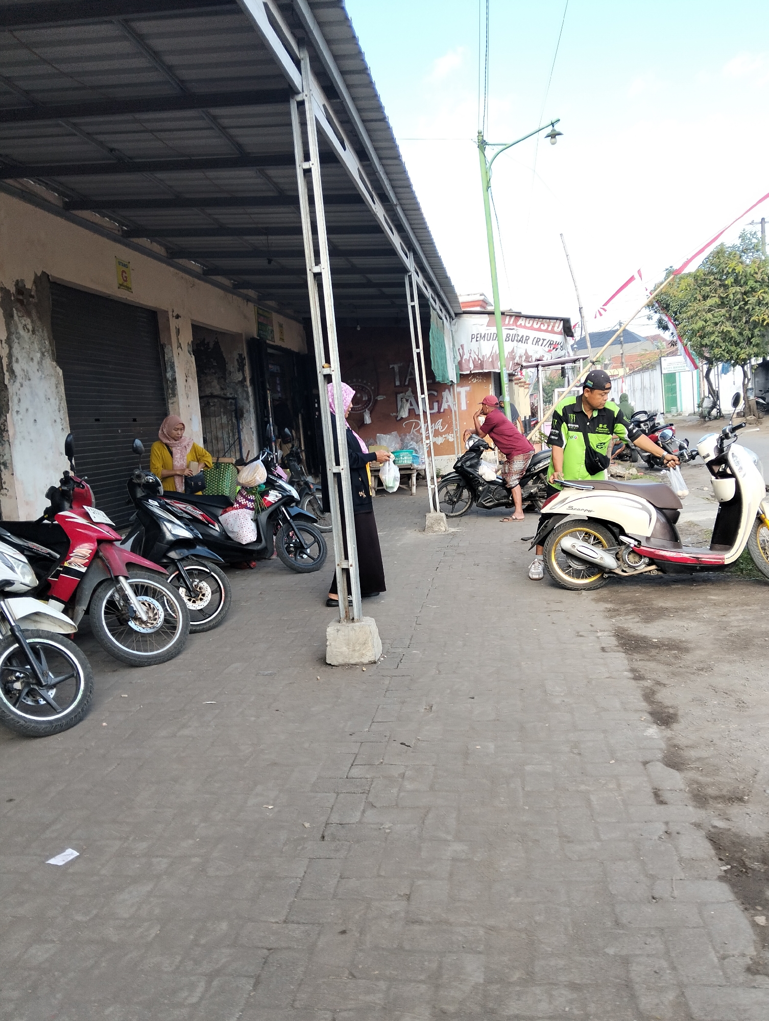 In Muncar, Indonesia, a street scene unfolds in front of a row of shops with metal roofs supported by metal poles. Several parked motorcycles, including a red and white one, a black one, and a cream and red scooter, line the paved sidewalk.
To the left, a woman wearing a yellow hijab and a light yellow top stands next to a black motorcycle, holding a bag. Another woman in a dark outfit and a pink hijab walks past the motorcycles.
In the center and right of the image, a man in a green shirt, black cap, and black fanny pack is interacting with a cream-colored scooter. A man in a maroon t-shirt and patterned shorts is standing near a black motorcycle.
A banner displaying "11 AGUSTUS PEMUD BUSAR (RT/RW)" is visible on a building in the background. The sky is bright and clear, with a green lamppost and some trees visible in the distance. The ground is a mix of paved interlocking bricks and dirt.