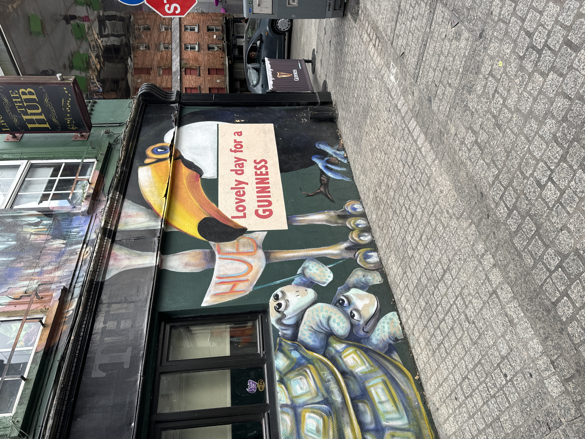 The image captures a mural on the side of a building, likely a pub or bar, in Waterford, Ireland. The mural features cartoonish turtles in the foreground and a toucan with a sign that reads "Lovely day for a GUINNESS." Above the mural, a sign for "THE HUB" is visible. The building is painted green and has windows with dark frames. The street in front of the building is paved with cobblestones. In the background, there are brick buildings and a dark-colored car. The overall impression is of a lively urban setting.