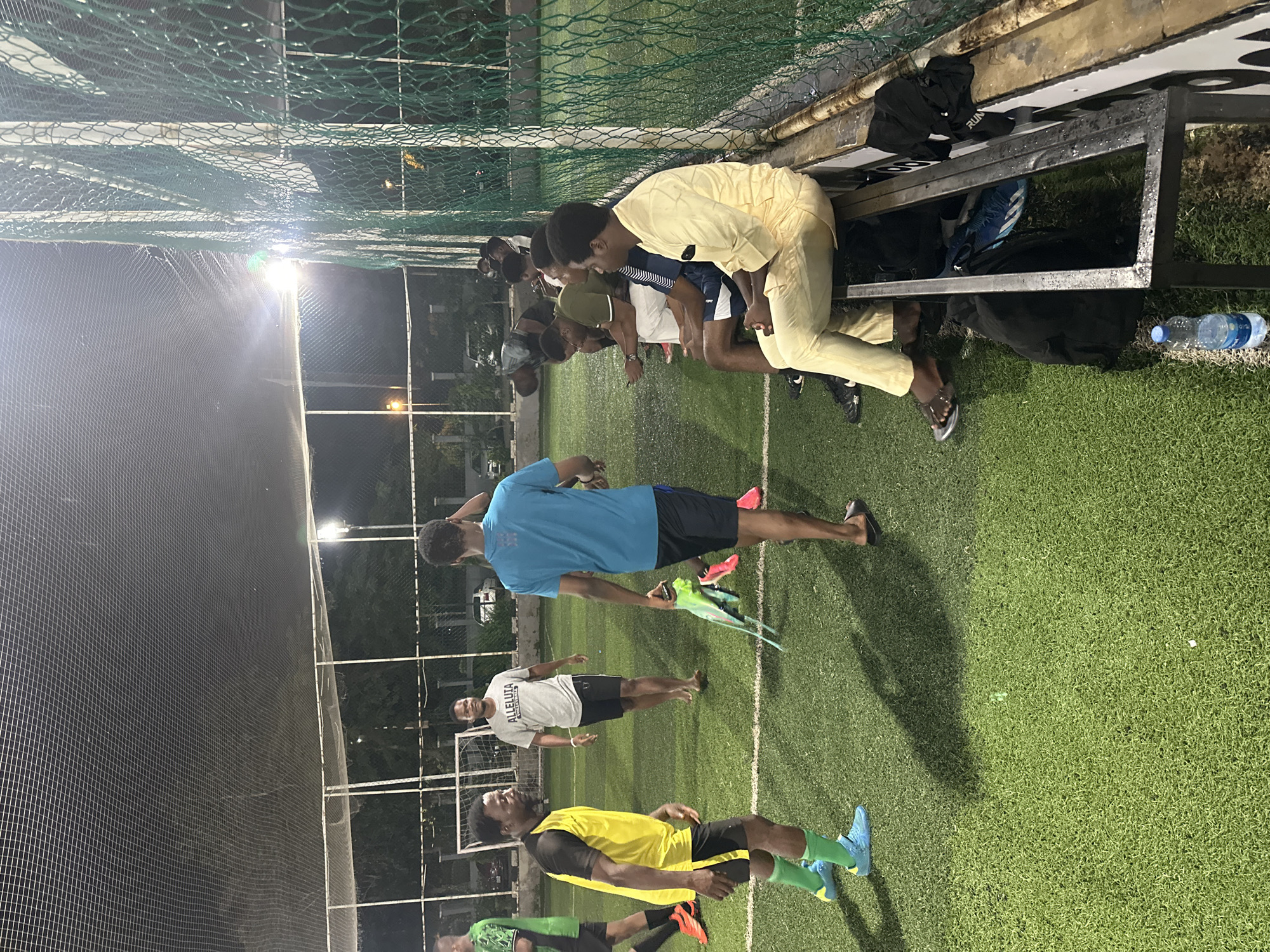 The image depicts a group of men on an artificial turf football pitch at night, possibly in Abuja, Nigeria. The scene is illuminated by artificial lights, casting long shadows across the green surface. The men are dressed in athletic wear, with some actively playing and others observing from the sidelines.
In the foreground, a man in a yellow and black jersey is in motion, his attention directed to his left. To his right, another man in a blue t-shirt carries a bundle of bright green sports equipment. In the background, a man wearing a grey t-shirt with "ALLELUIA VOLLEYBALL" printed on it stands looking towards the players.
A bench runs along the right side of the pitch, where several men are seated, some looking at their phones. The pitch is enclosed by a green net and metal fencing. A water bottle is visible on the ground near the bench. The overall atmosphere suggests a casual game or practice session.