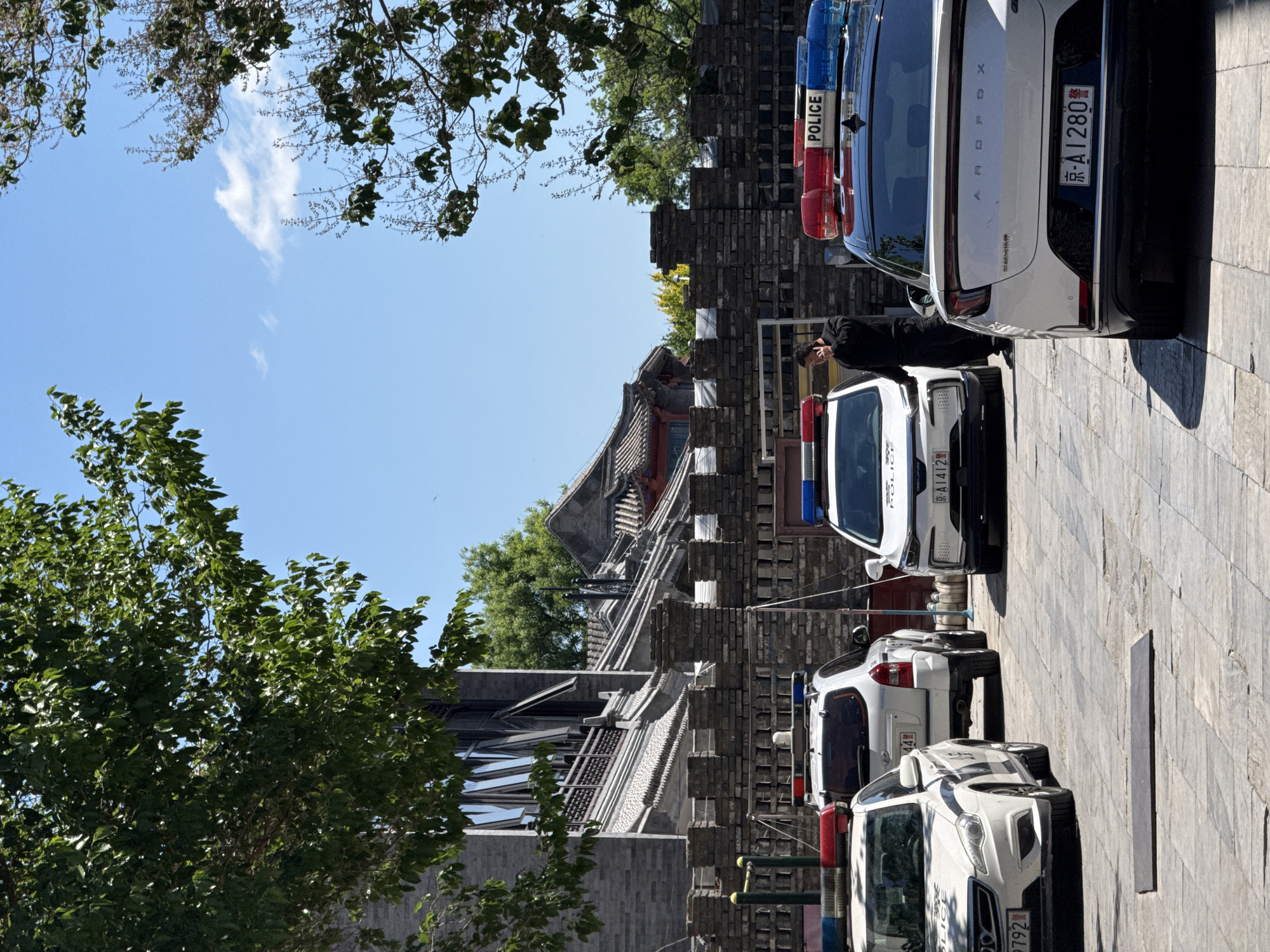 The image shows a street scene in Dongcheng District, China, with several police cars parked alongside a traditional building. The vehicles, marked with "POLICE" in English, face away from the camera. An individual in dark clothing stands near one of the cars, possibly on a phone. The architecture features traditional Chinese rooflines and brickwork. Trees frame the top and left sides of the shot, with a clear blue sky visible above. The ground is paved with stone slabs.