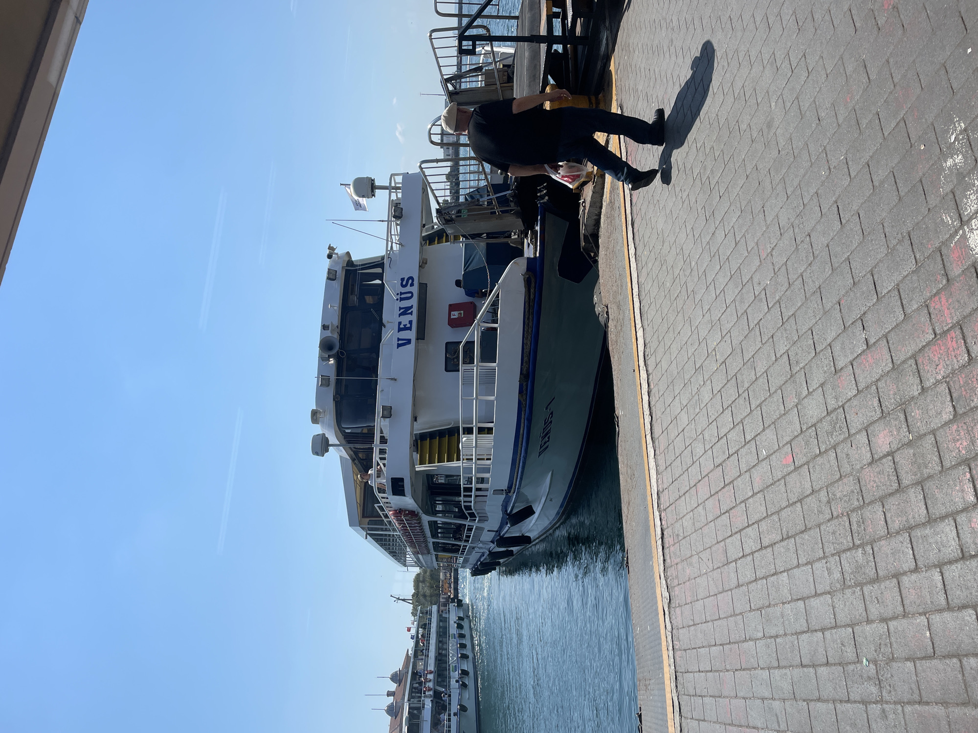 A ferry named VENUS-1 is docked at a pier in Kadıköy, Turkey. The ferry is white with blue trim and has multiple decks. A man in a dark t-shirt, blue jeans, and a light-colored flat cap is walking away from the ferry on a paved walkway. He is carrying a white plastic bag with red lettering. In the background, another ferry is visible in the water, and a building with dome-shaped roofs is on the shoreline. The sky is clear and blue, and the water is calm.