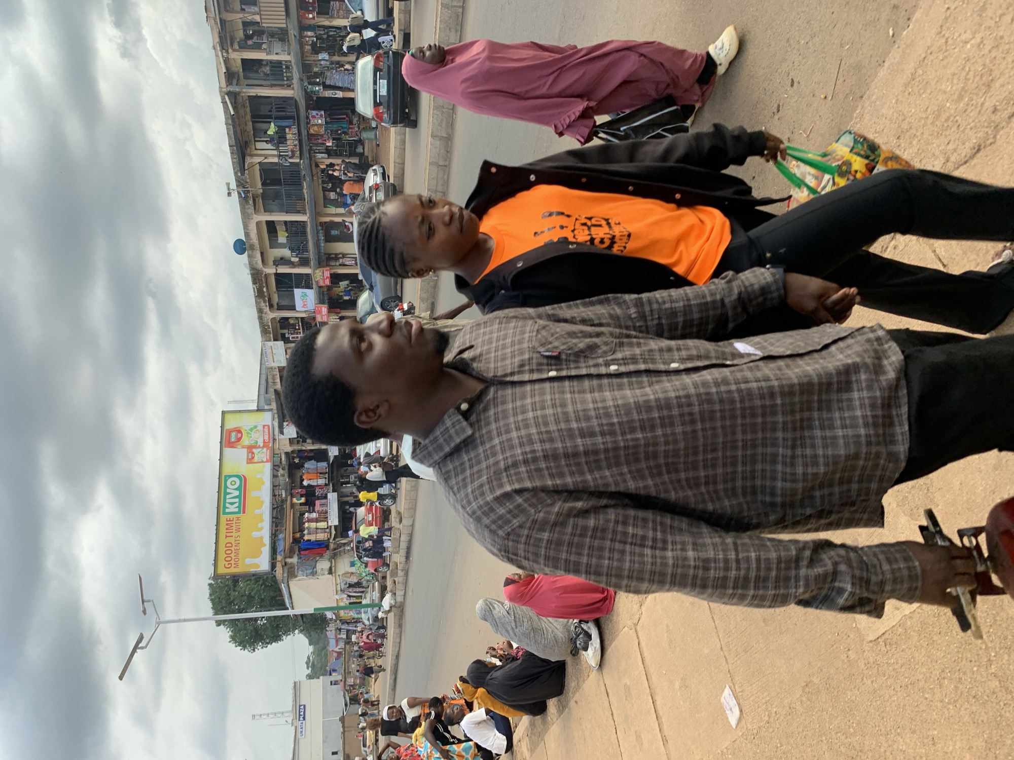 The image depicts a bustling street scene in Jos, Nigeria, likely during the daytime given the lighting. The perspective is low, close to the ground, with the foreground occupied by the shoulders and head of a man wearing a plaid shirt. To his right, a young woman in an orange t-shirt and black jacket is also in the foreground, looking to her right. They appear to be sitting or standing together on a sidewalk.
In the background, a market or commercial area lines the street. Shops with goods displayed are visible, and several people are walking and milling about. A prominent billboard advertises "GOOD TIME MOMENTS WITH KIVO" with images of juice. Cars and motorcycles are present, suggesting ongoing traffic. A few individuals are seated on the ground by the sidewalk, some wearing headscarves. The overall atmosphere is one of everyday activity in a busy urban environment. The sky appears overcast, indicating a cloudy day.