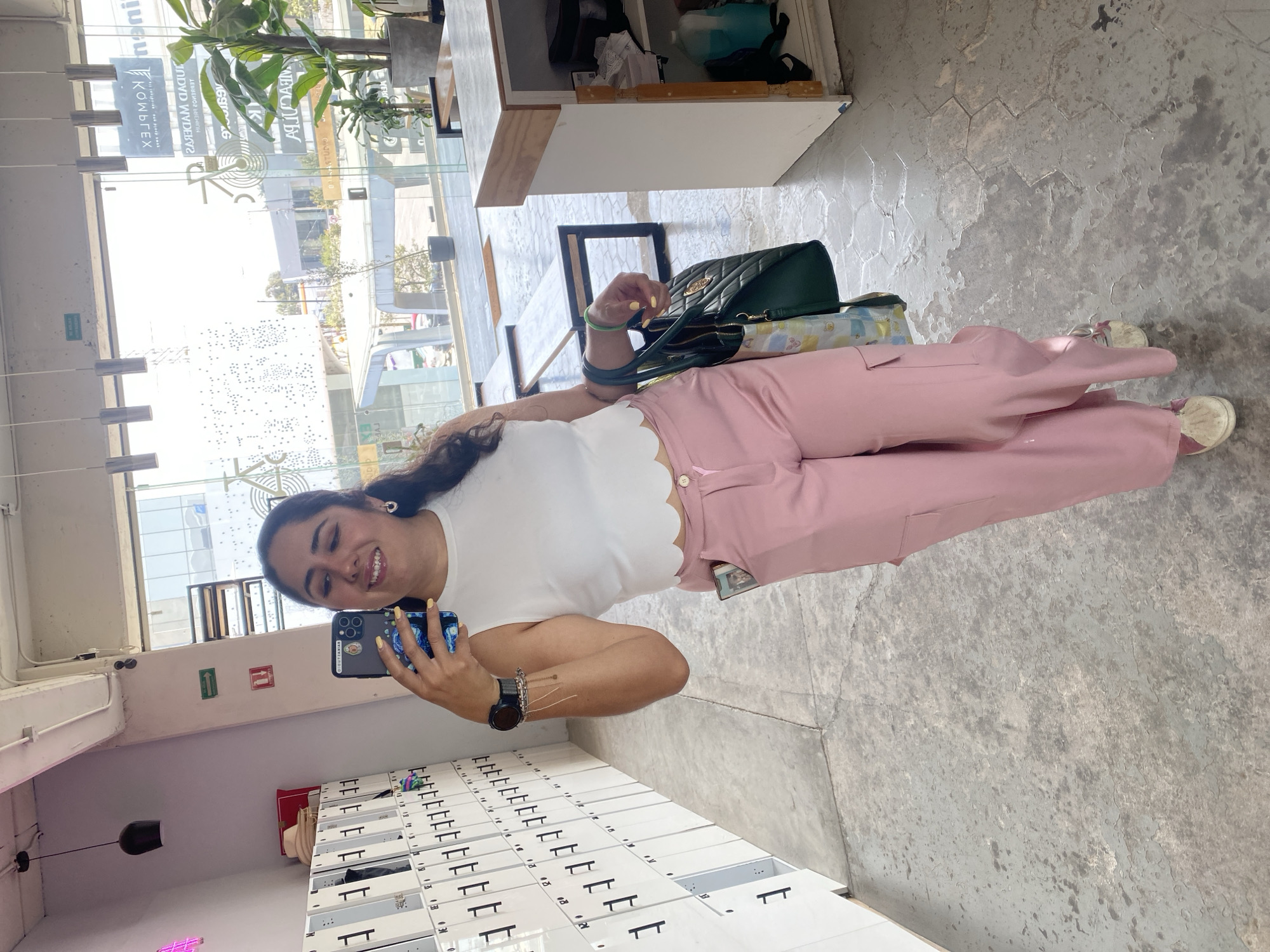 A smiling woman stands in a space with white lockers against a concrete wall, holding a smartphone and taking a selfie. She wears a white top, pink pants, and white shoes. A green purse rests on the floor beside her. Natural light enters from a large window showing a city scene with buildings and signs, potentially in Puebla, Mexico. The interior has a modern, minimalist aesthetic.